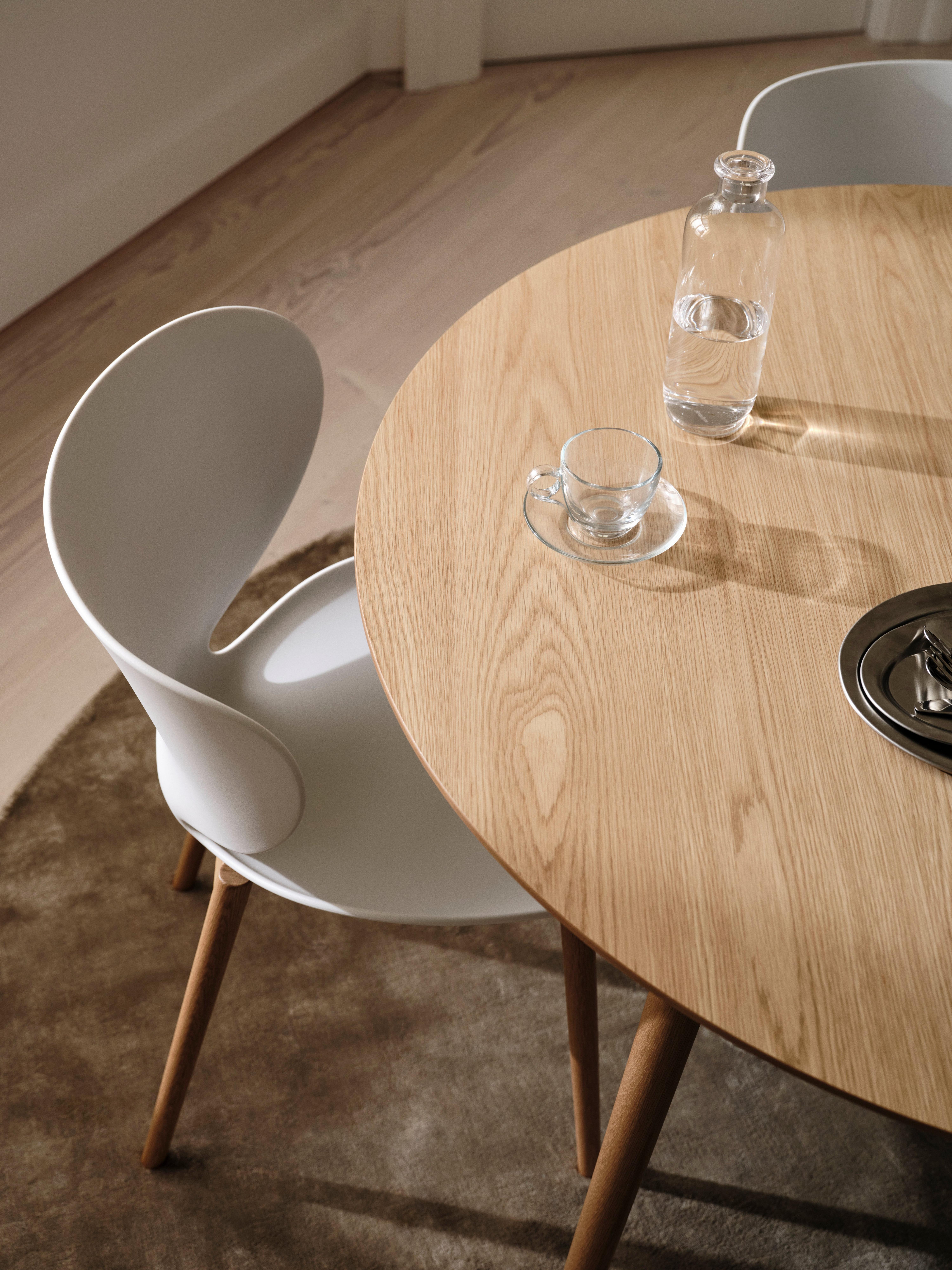 Round wooden table with glass bottle, cup, saucer, and white chair on a rug.