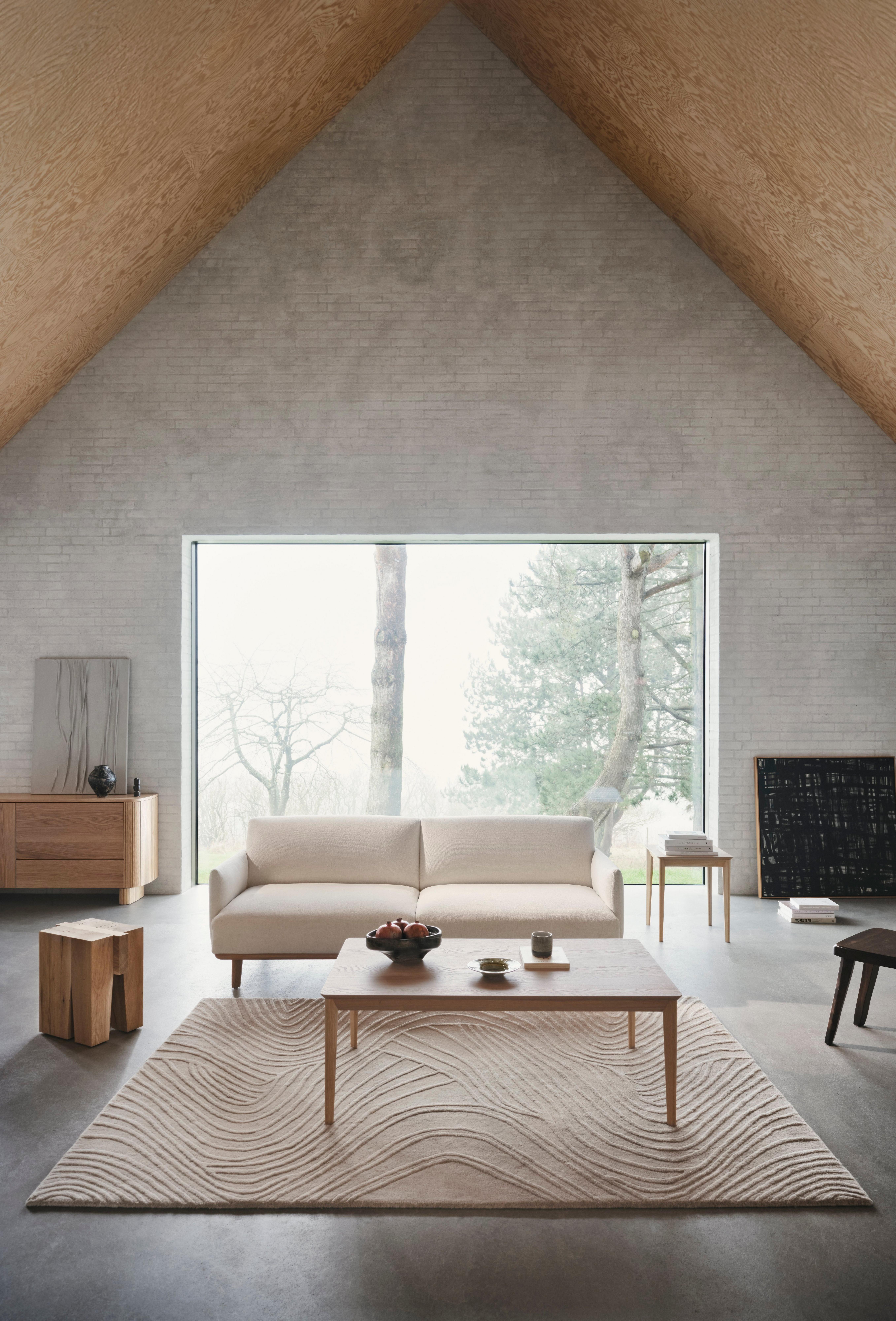 Minimalist living room with a light sofa, wood coffee table, and textured rug, set against a tall window and angled wood ceiling.