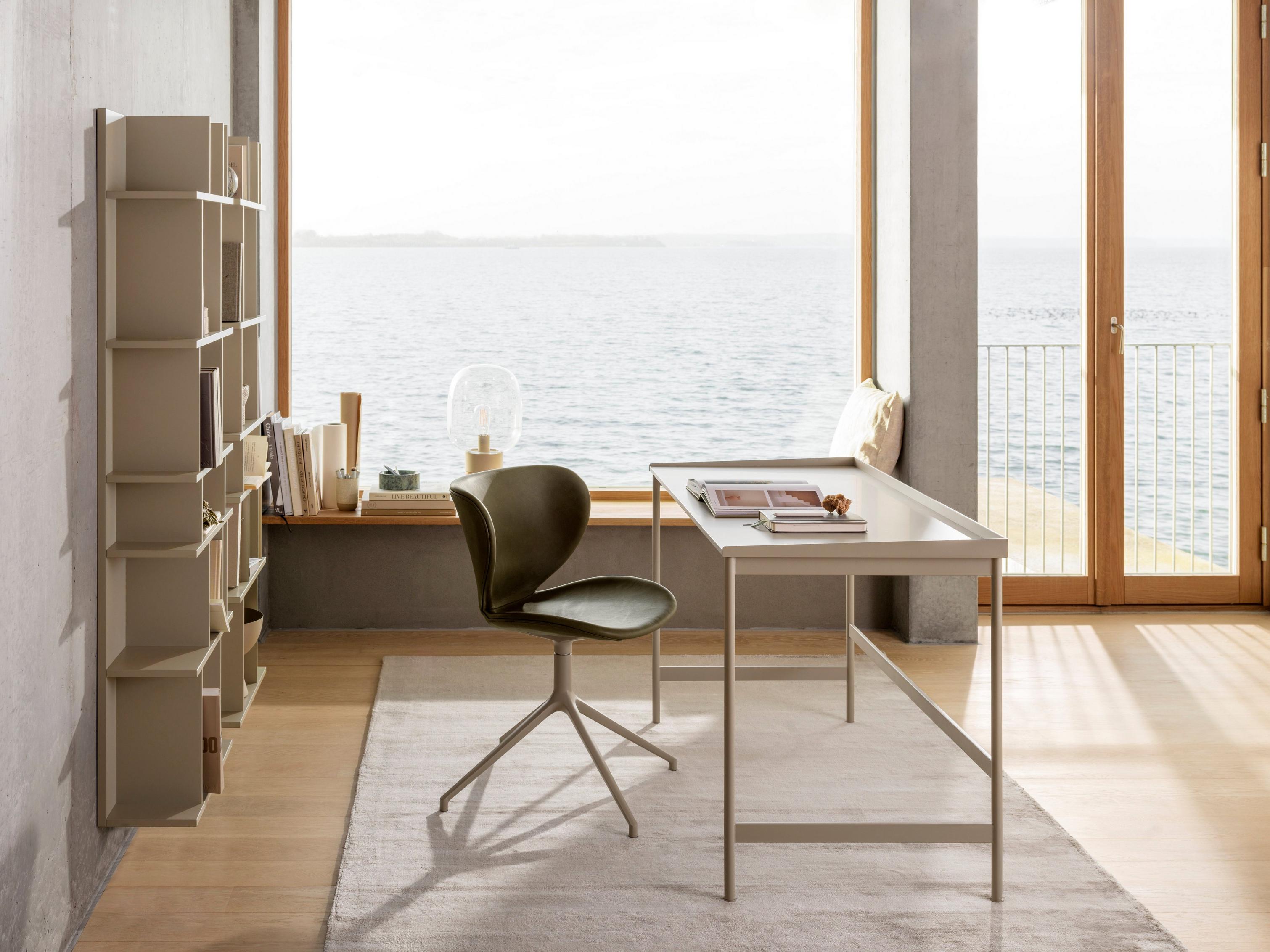 Seaside home office with a white desk, green chair, and shelving, with large windows and ocean view.