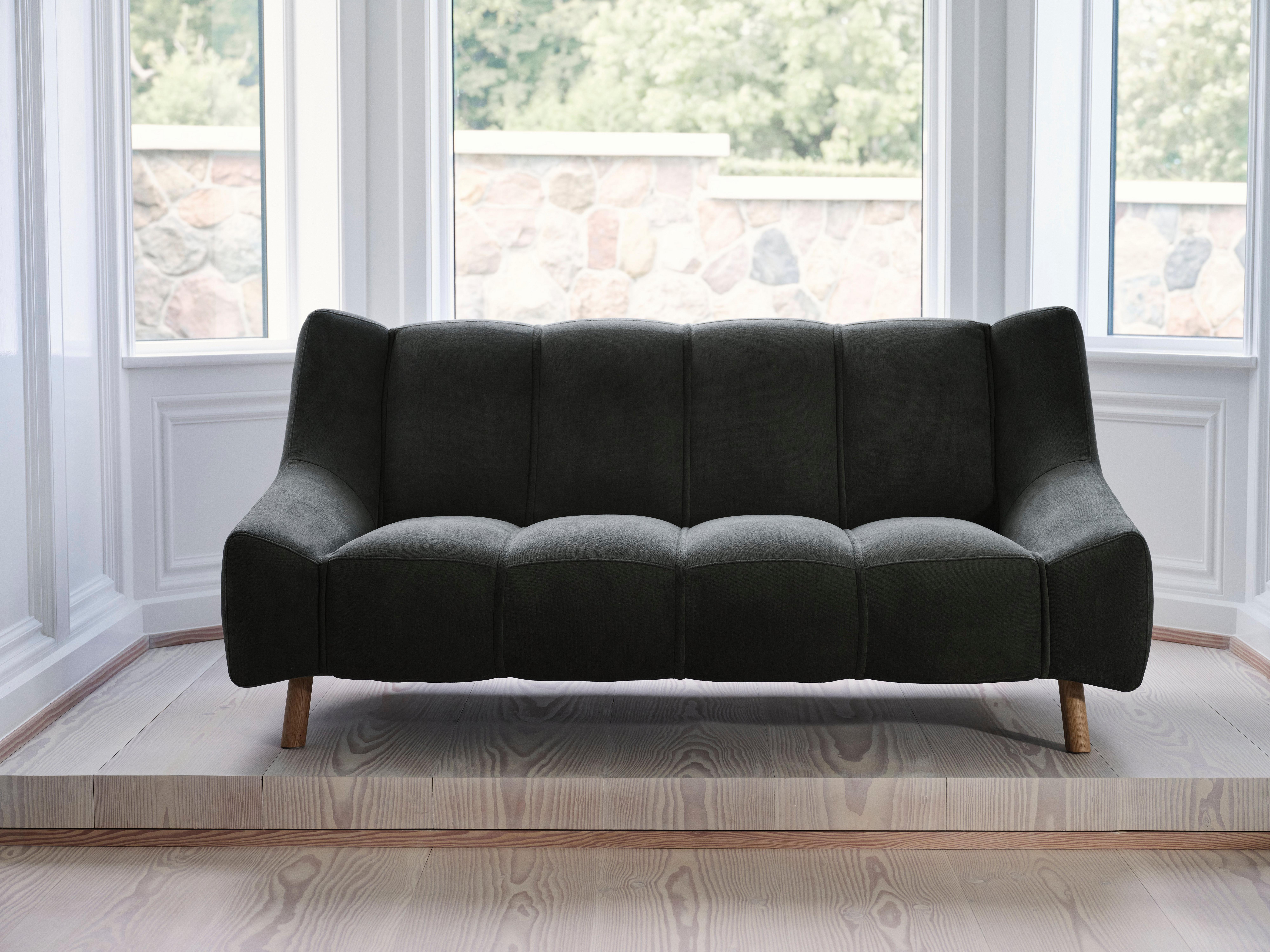 Tumma sohva puisilla jaloilla ikkunaseinän edessä.