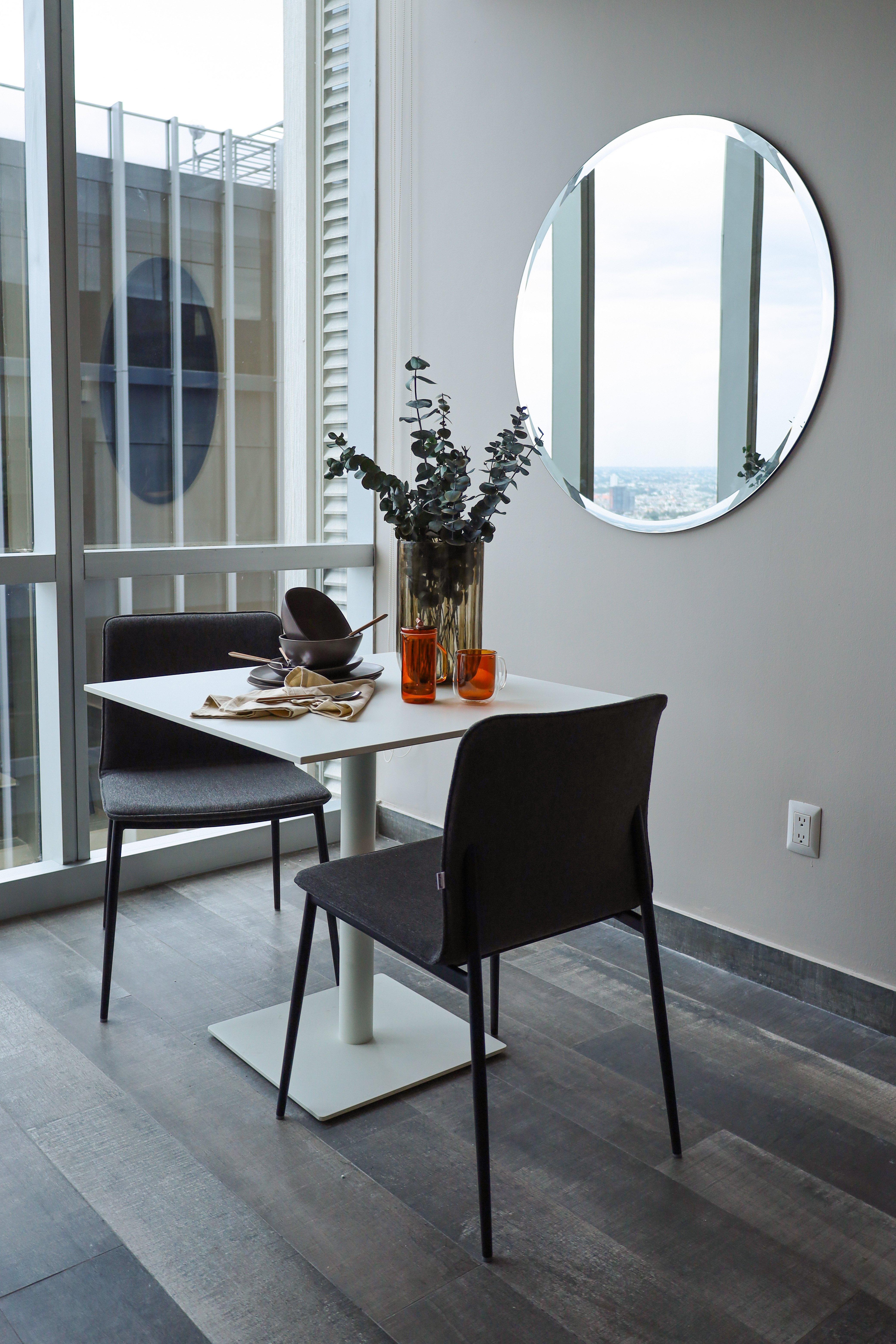 Modern dining area with two chairs, a round mirror, and a table set with crockery and orange glasses.