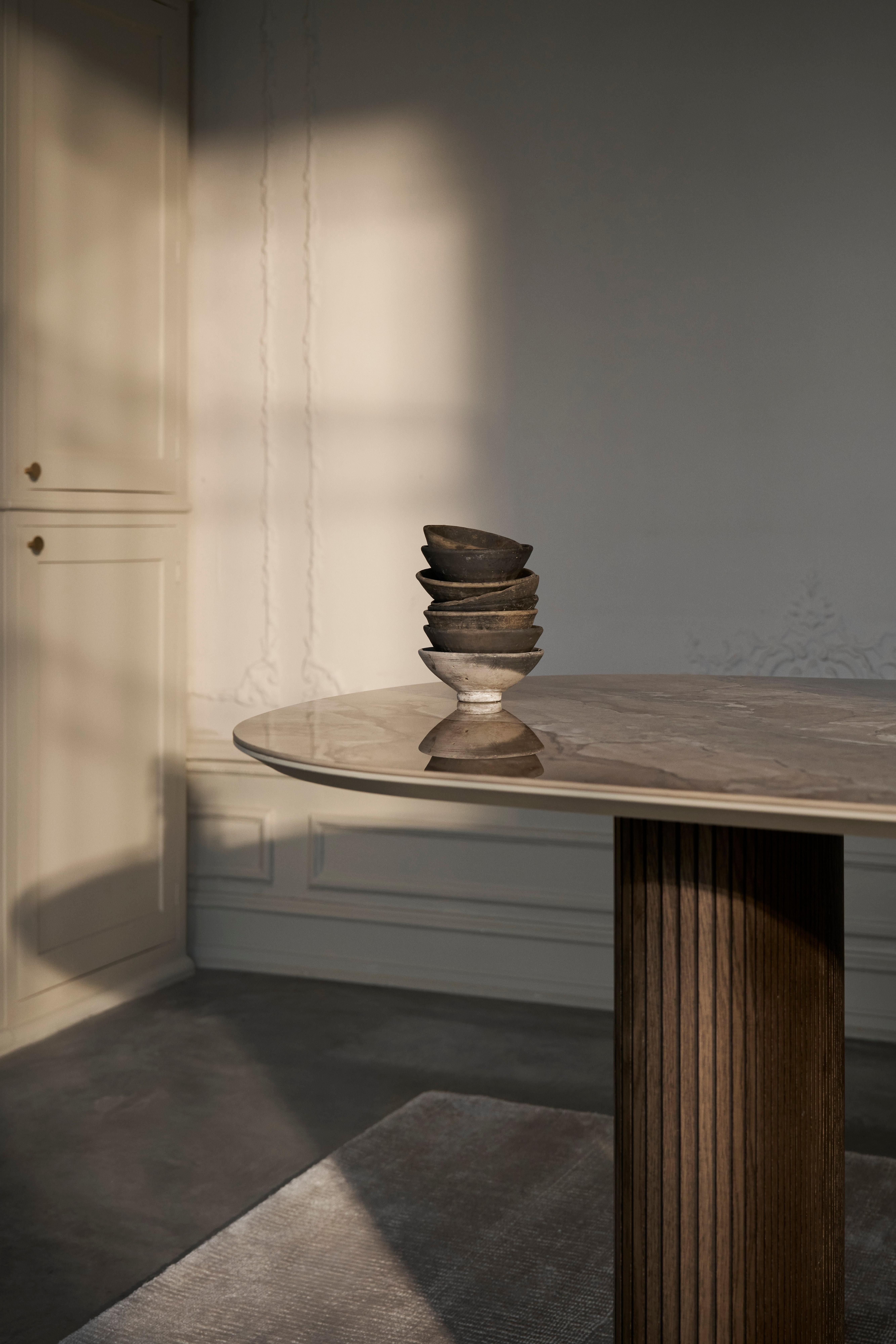 Santiago dining table in brown ceramic with fluted wooden base, placed on a stone grey Simple rug in a softly lit room.