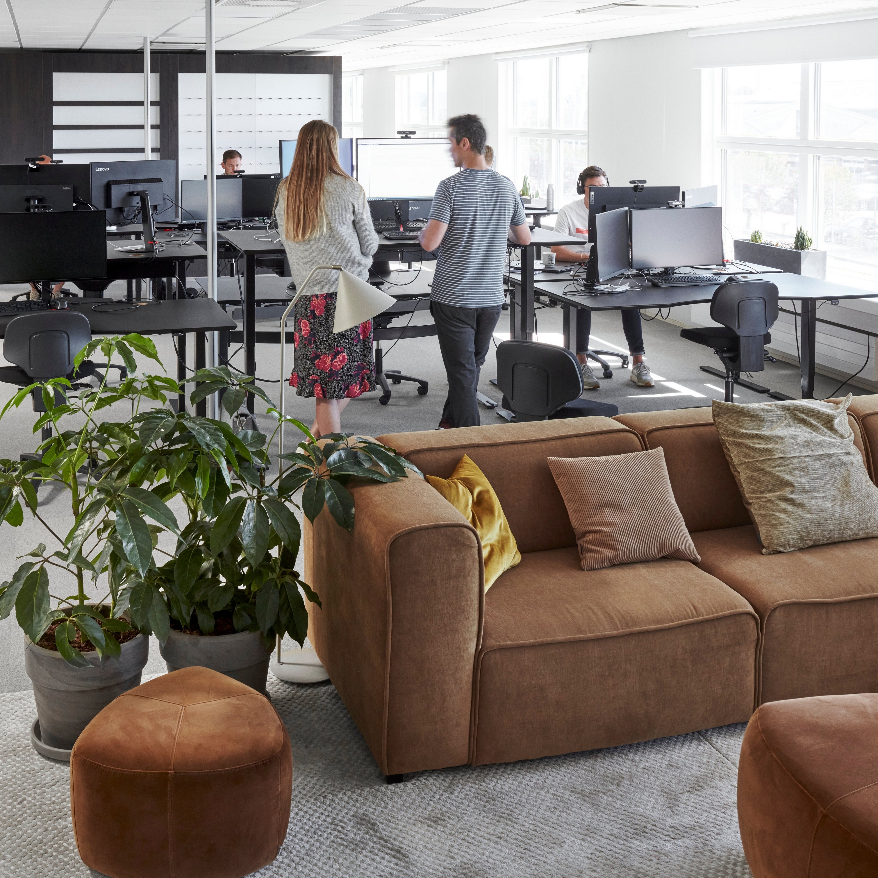 Espacio de oficina moderno en el corporativo de BoConcept con empleados, computadoras, plantas y un sofá Carmo marrón.