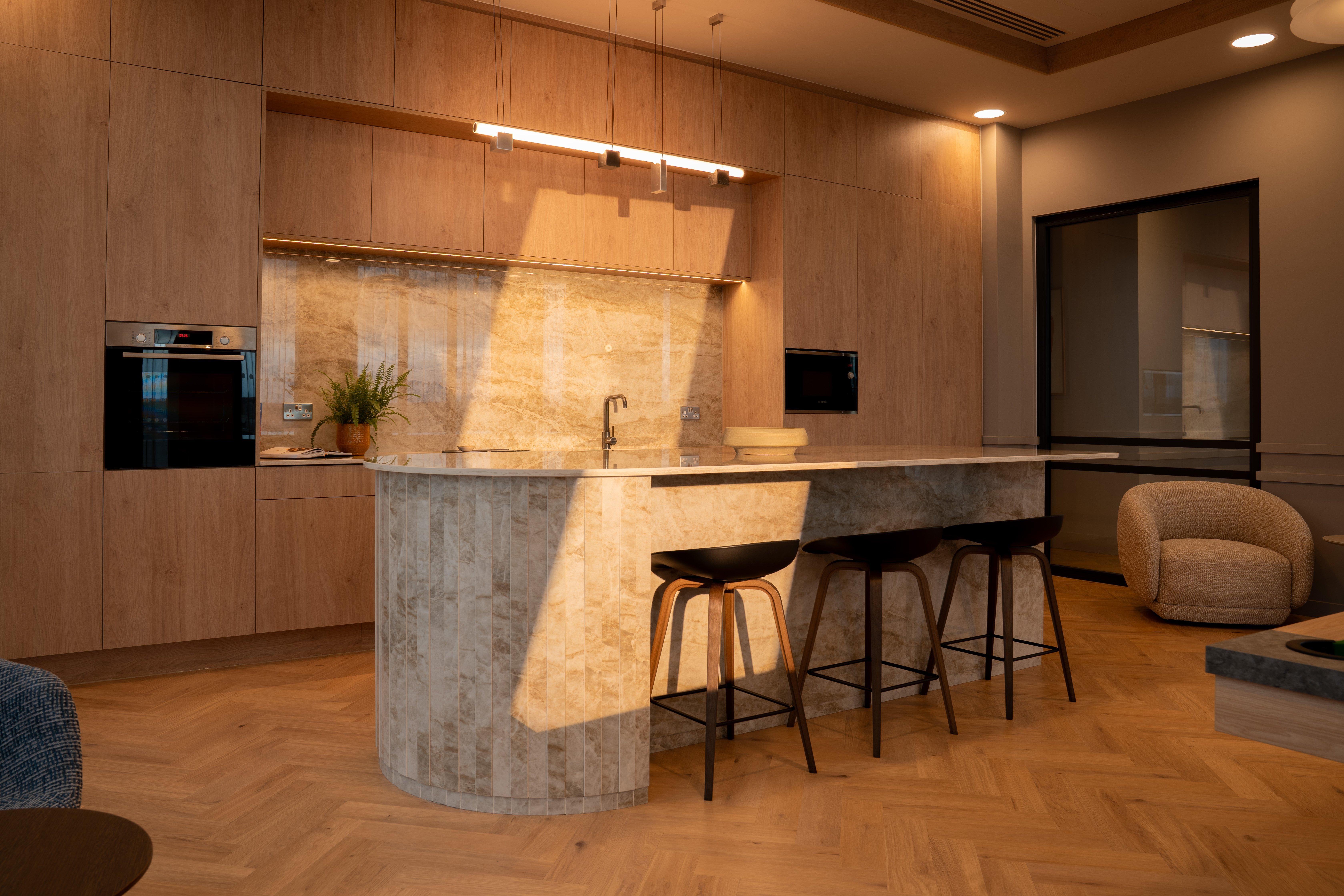 Cocina moderna con isla de mármol, tres taburetes altos y armarios de madera.