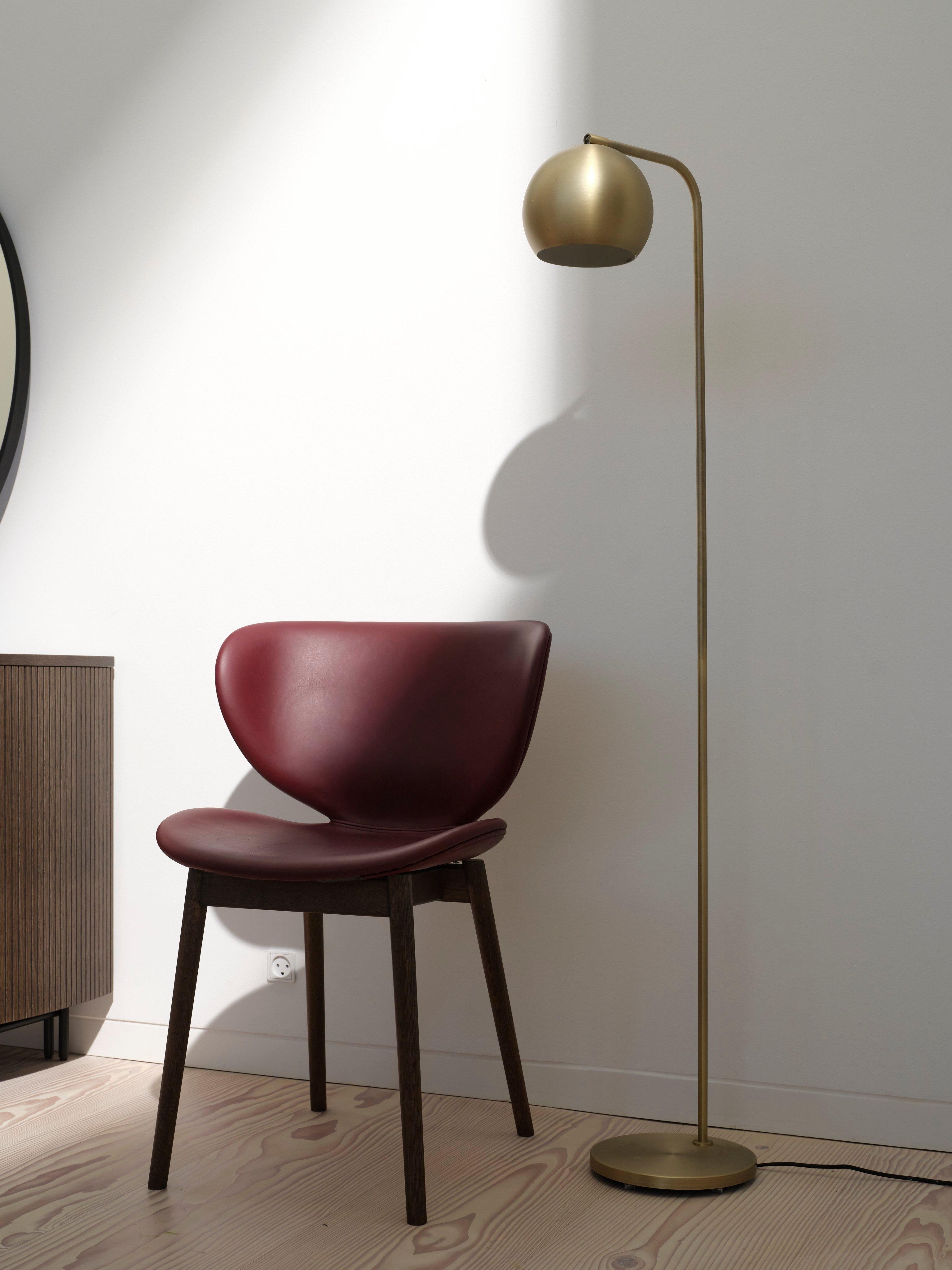 Kožená stolička Hamilton vo vínovo červenej farbe vedľa zlatej stojanovej lampy pred bielou stenou s čiastočne viditeľným dreveným príborníkom.
