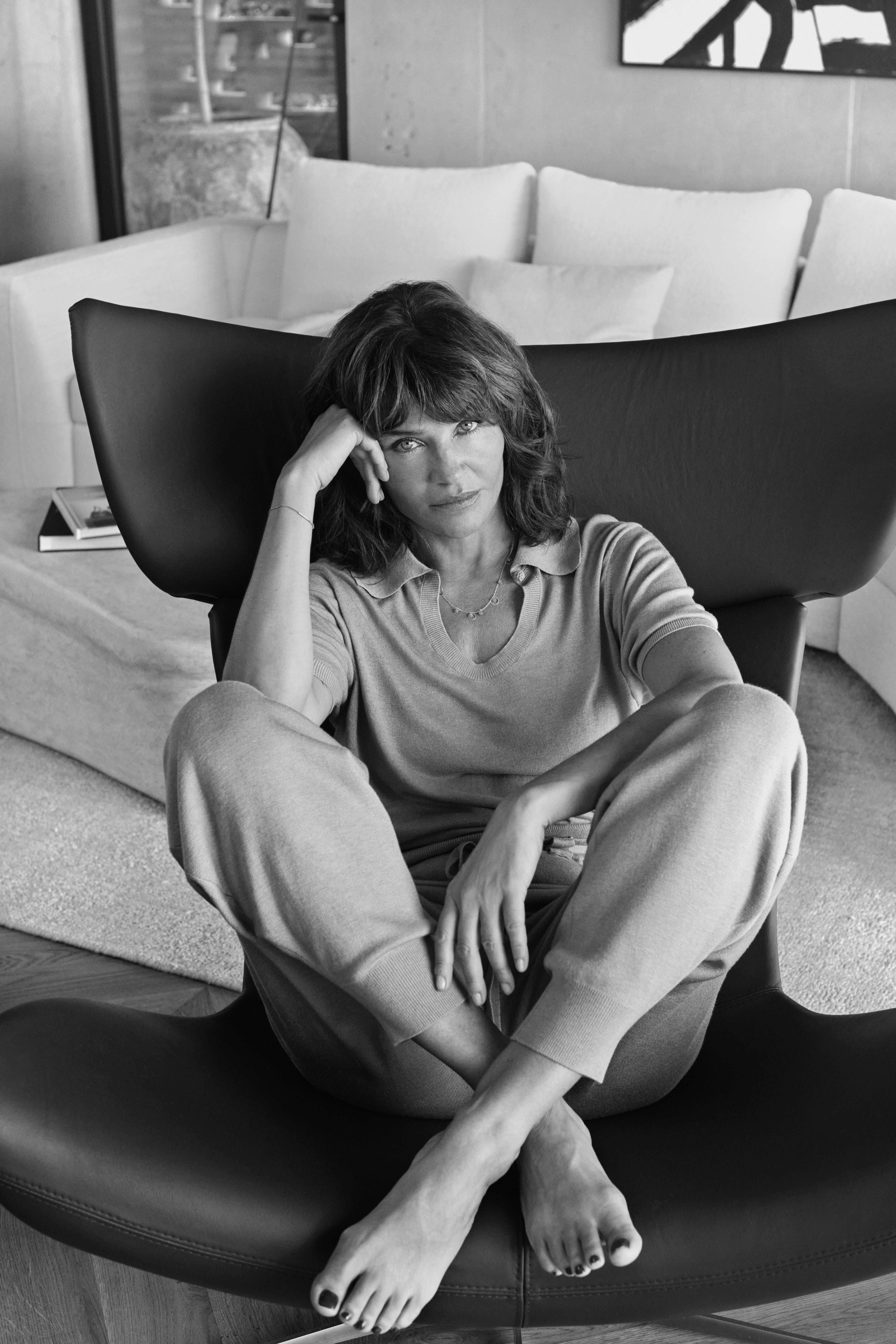 Black and white image of a Helena Christensen seated on a black Imola armchair, dressed in casual clothing, with a neutral interior background.