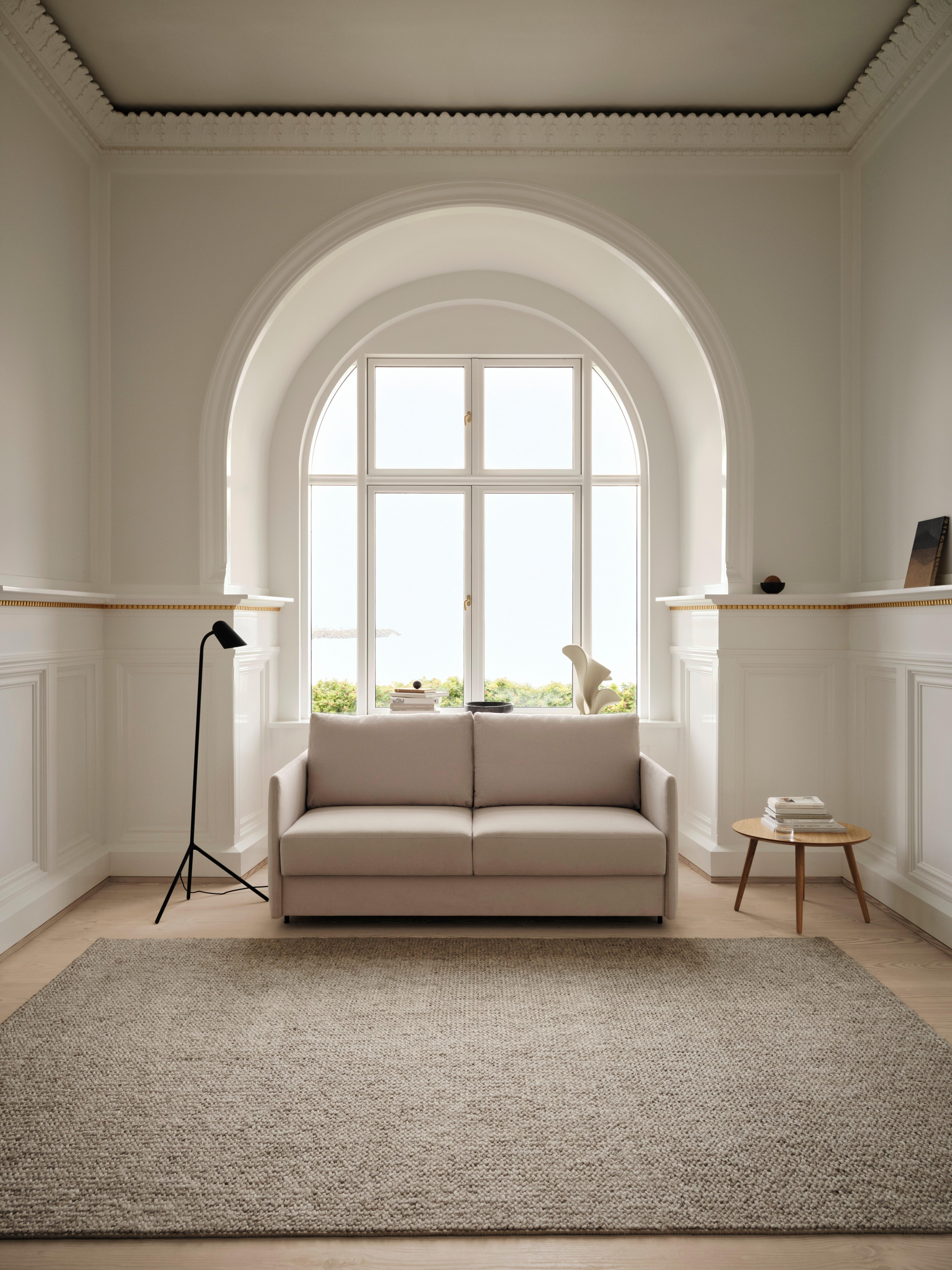Lyst stue med beige sofa, gulvlampe, og lille bord ved stort, buet vindue. Tæppe på gulvet.