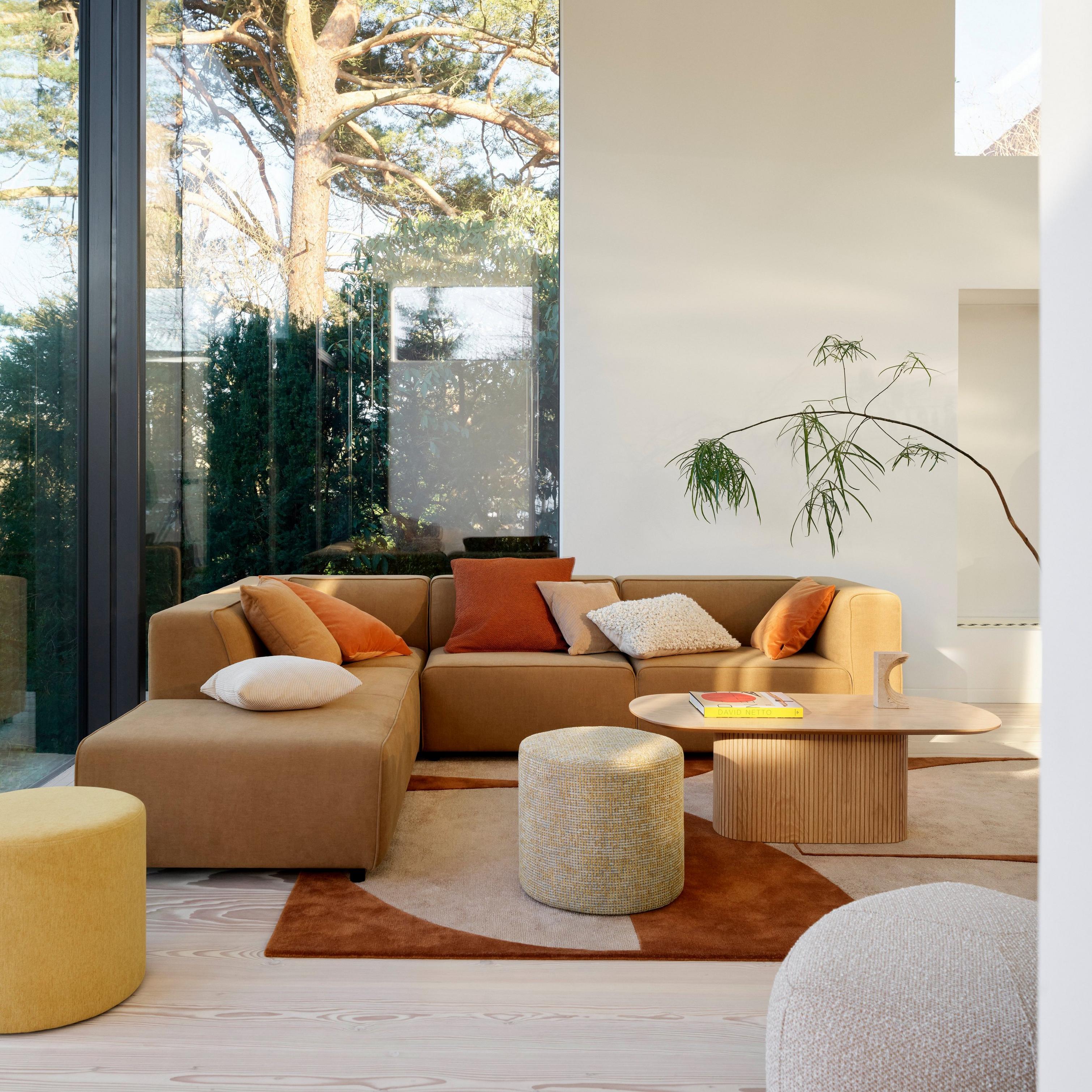 Cozy living room with a sectional sofa, various pillows, round Eden footstools, a Santiago coffee table, large windows, and a view of trees.