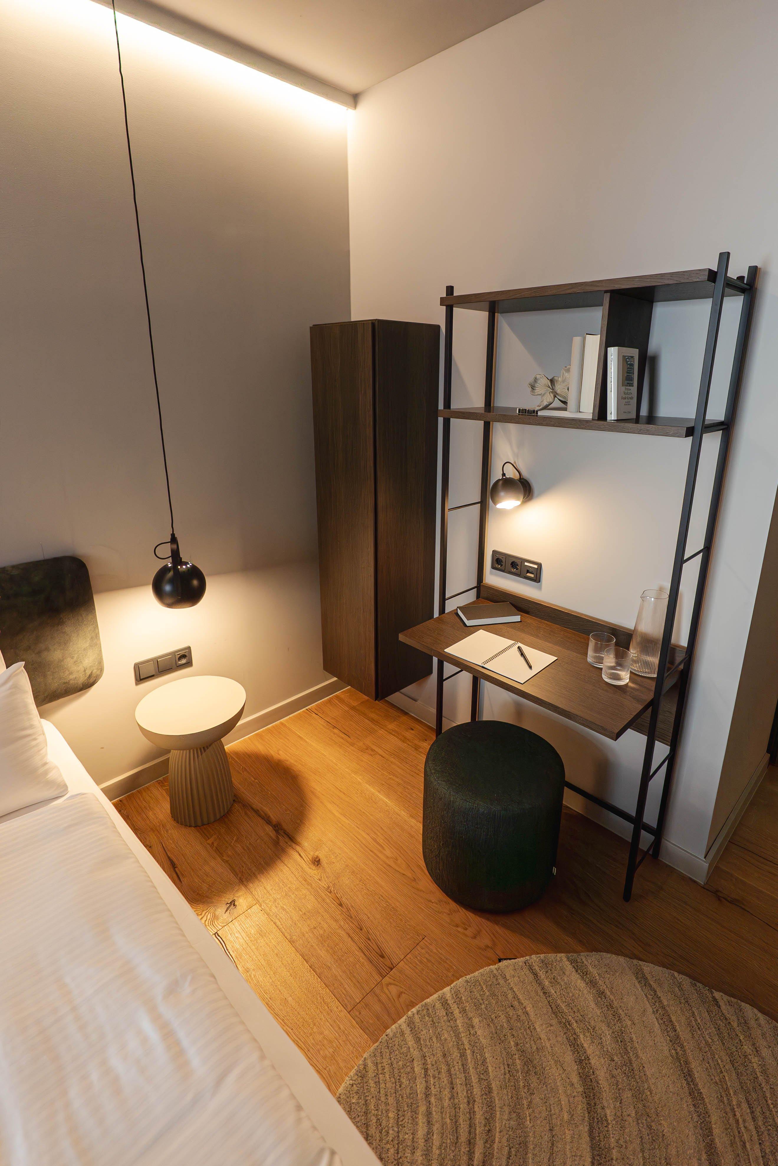 Bureau moderne avec étagères, lampe, carnet et carafe sur sol en bois. Tabouret et table d'appoint présents.