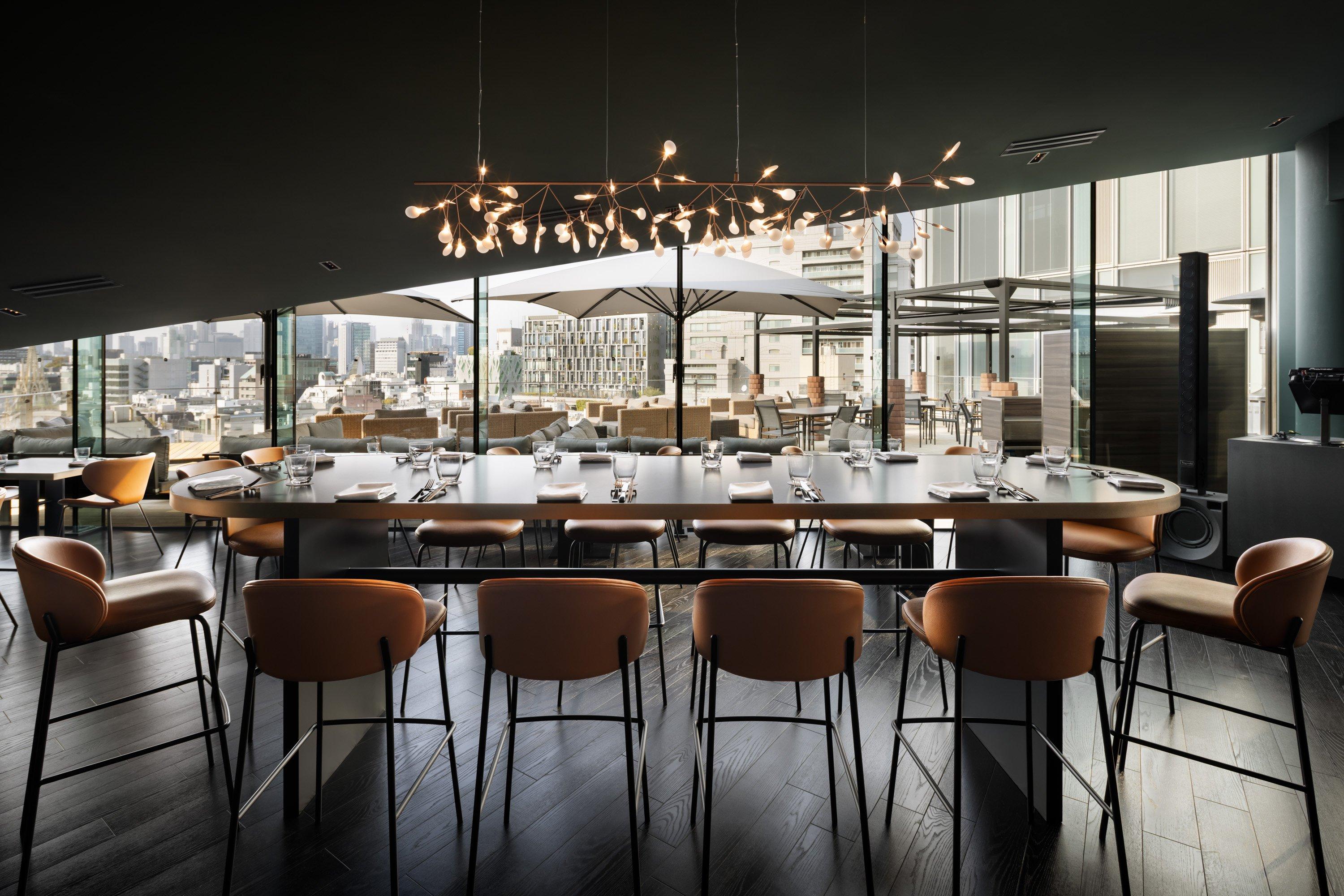 Restaurante moderno con mesa grande, sillas naranjas, lámpara decorativa y vista a la ciudad.