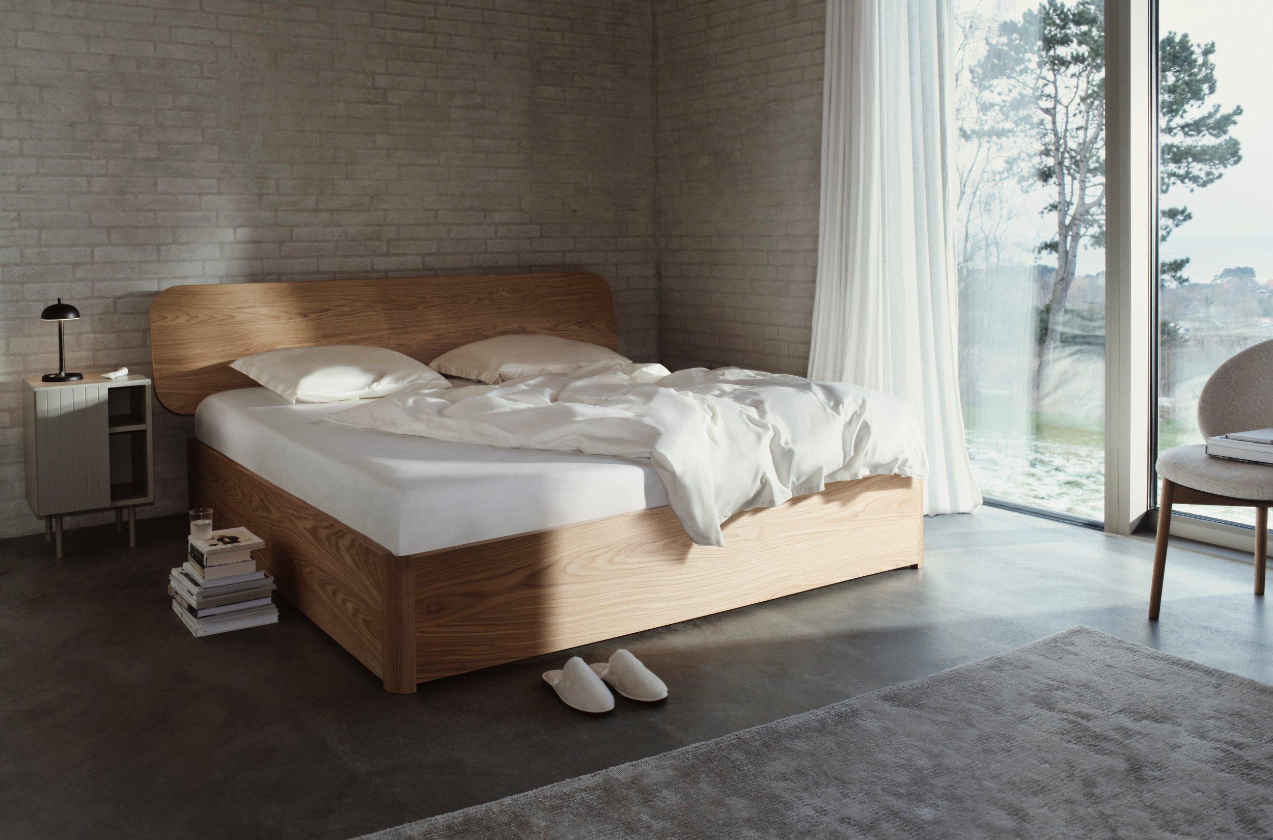 Minimalist bedroom with a wood-framed bed, white bedding, grey rug, and floor-to-ceiling window with sheer curtains and a scenic view.