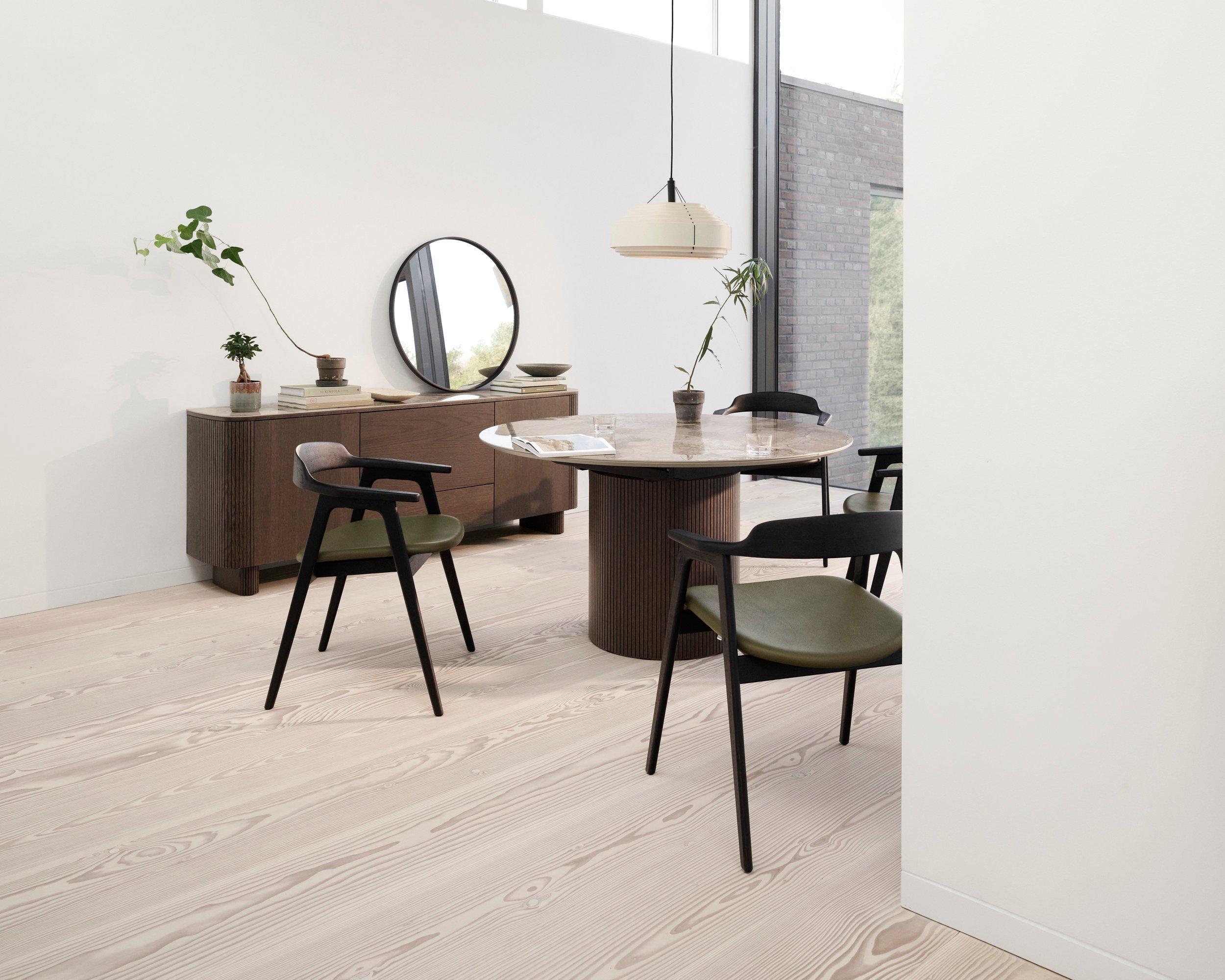 Minimalist dining room featuring a Santiago extendable dining table with a marble top, Santiago sideboard, and Seoul dining chairs with green cushions. Decor includes a Bubble vase, Ribbon vase, and a Paddle pendant light. The room has a round mirror on the sideboard and large windows.