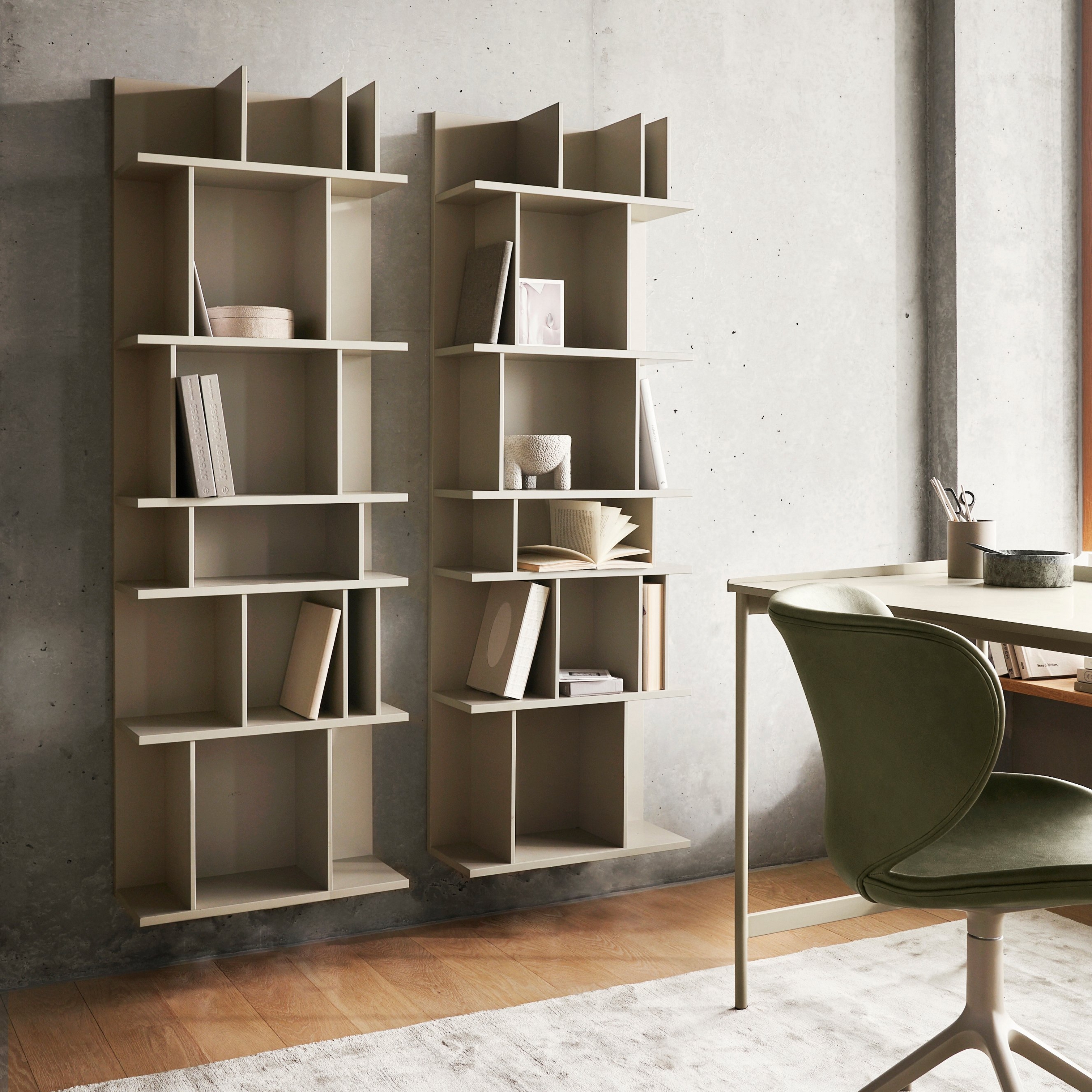 Asymmetrical wooden shelves with books, next to a desk and chair in a room with concrete walls.