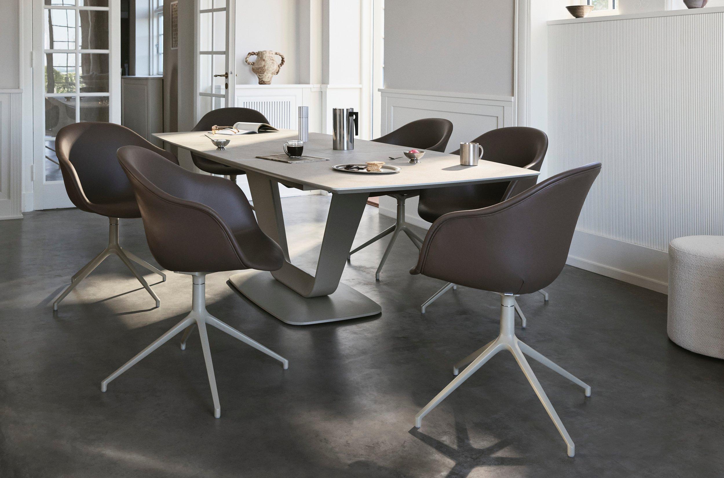 Modernes Esszimmer mit sechs Drehstühlen um einen quadratischen Tisch auf dunklem Boden in einem hellen Raum mit weissen Wänden und Fenstern.