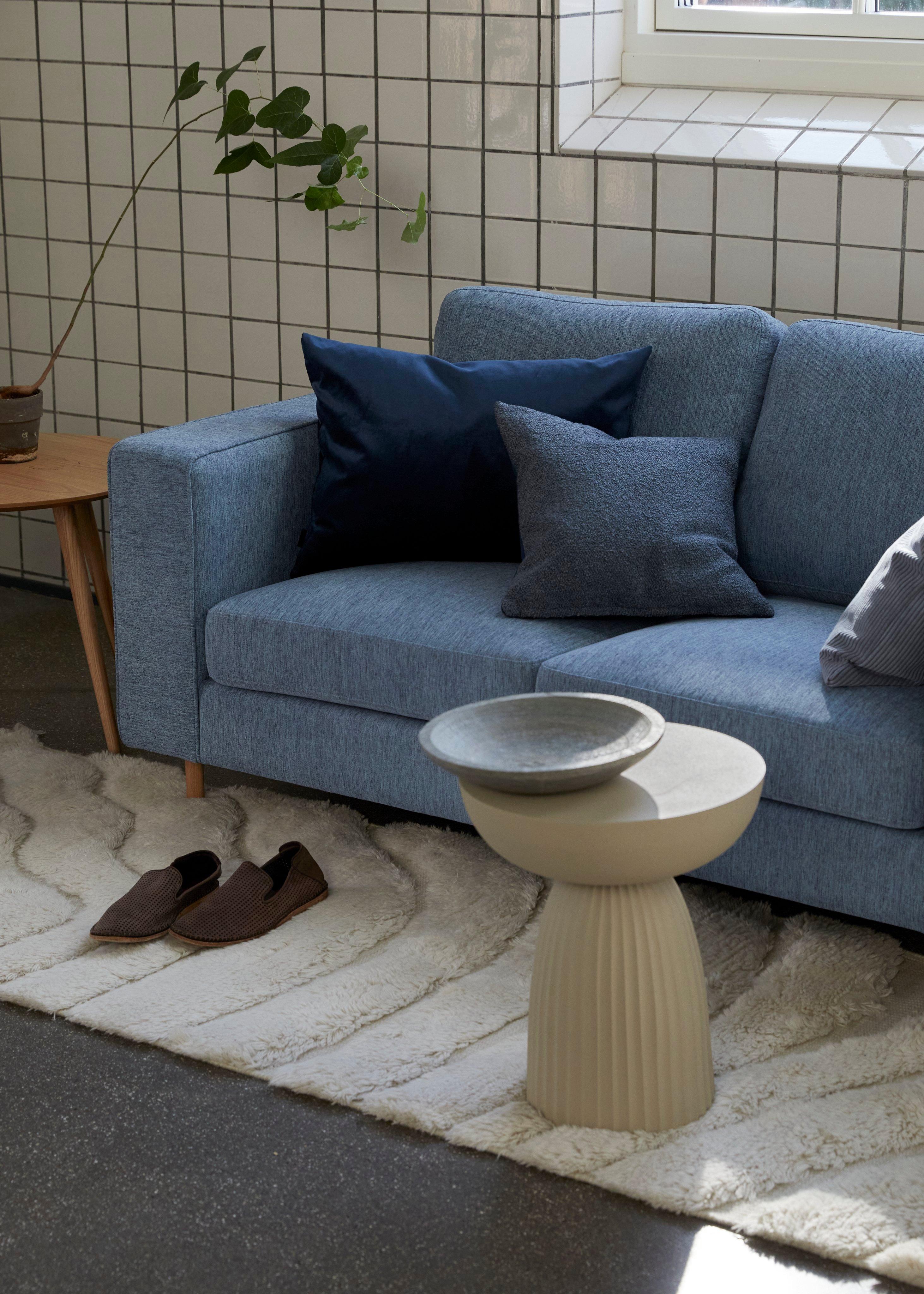 Living room with a blue Indivi sofa, blue and gray pillows, white textured rug, beige Expoe side table, potted plant, and slippers on the floor.