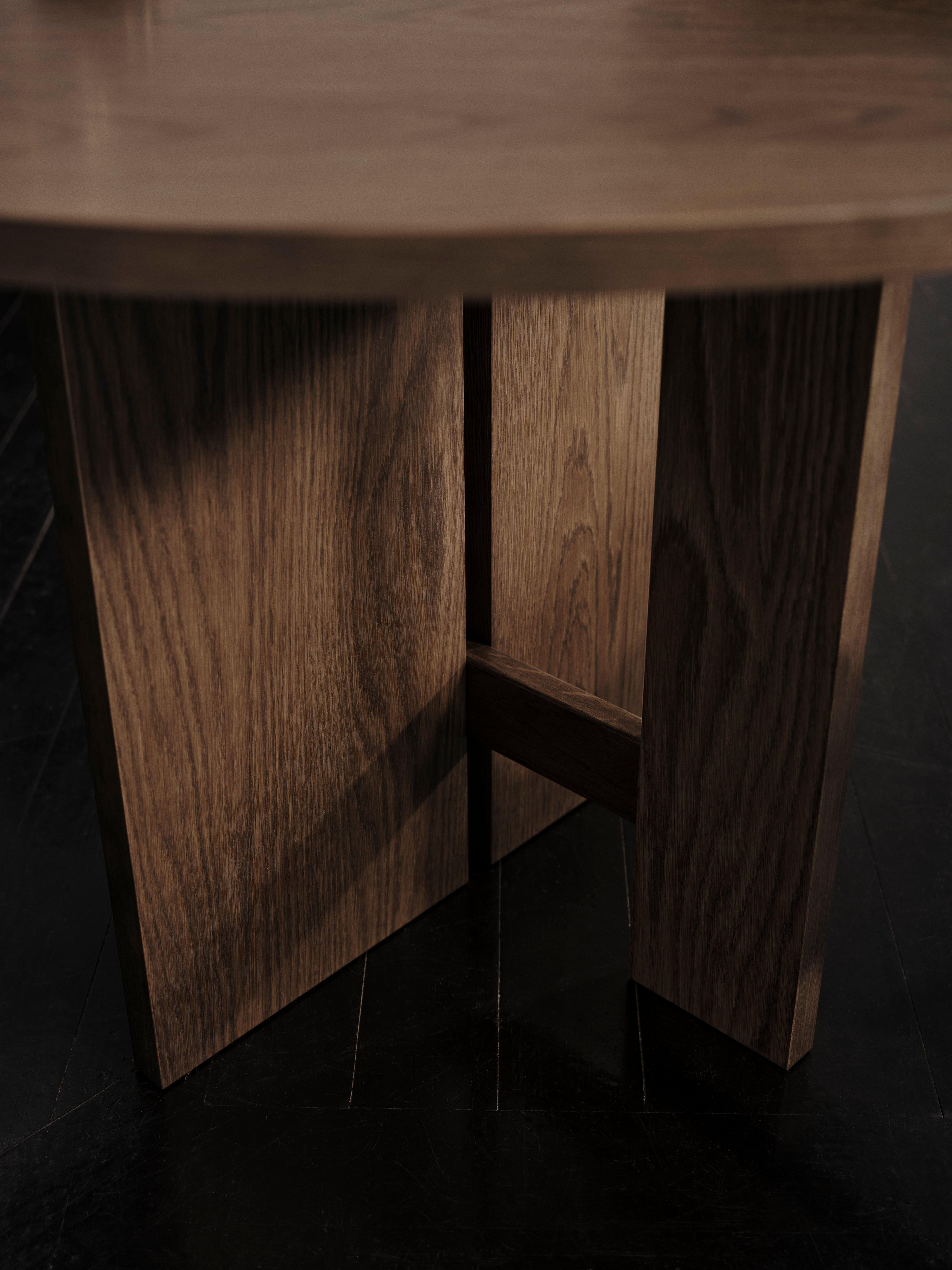 Wooden table with round top and sturdy, intersecting legs on a dark floor.