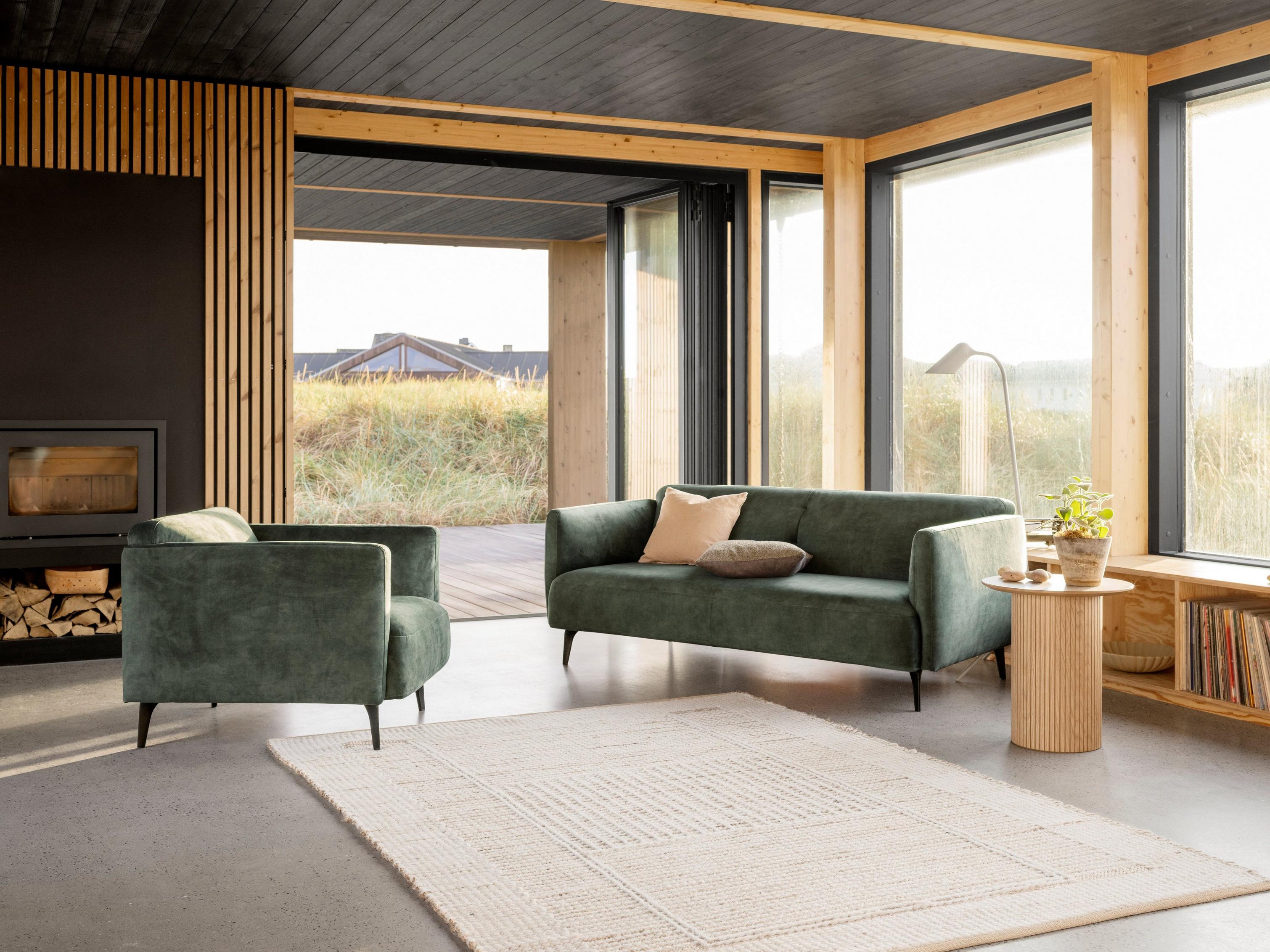 Den elegante Modena sofa og den matchende lænestol i støvet grønt Ravello stof står ved siden af pejsen i denne varme stue.