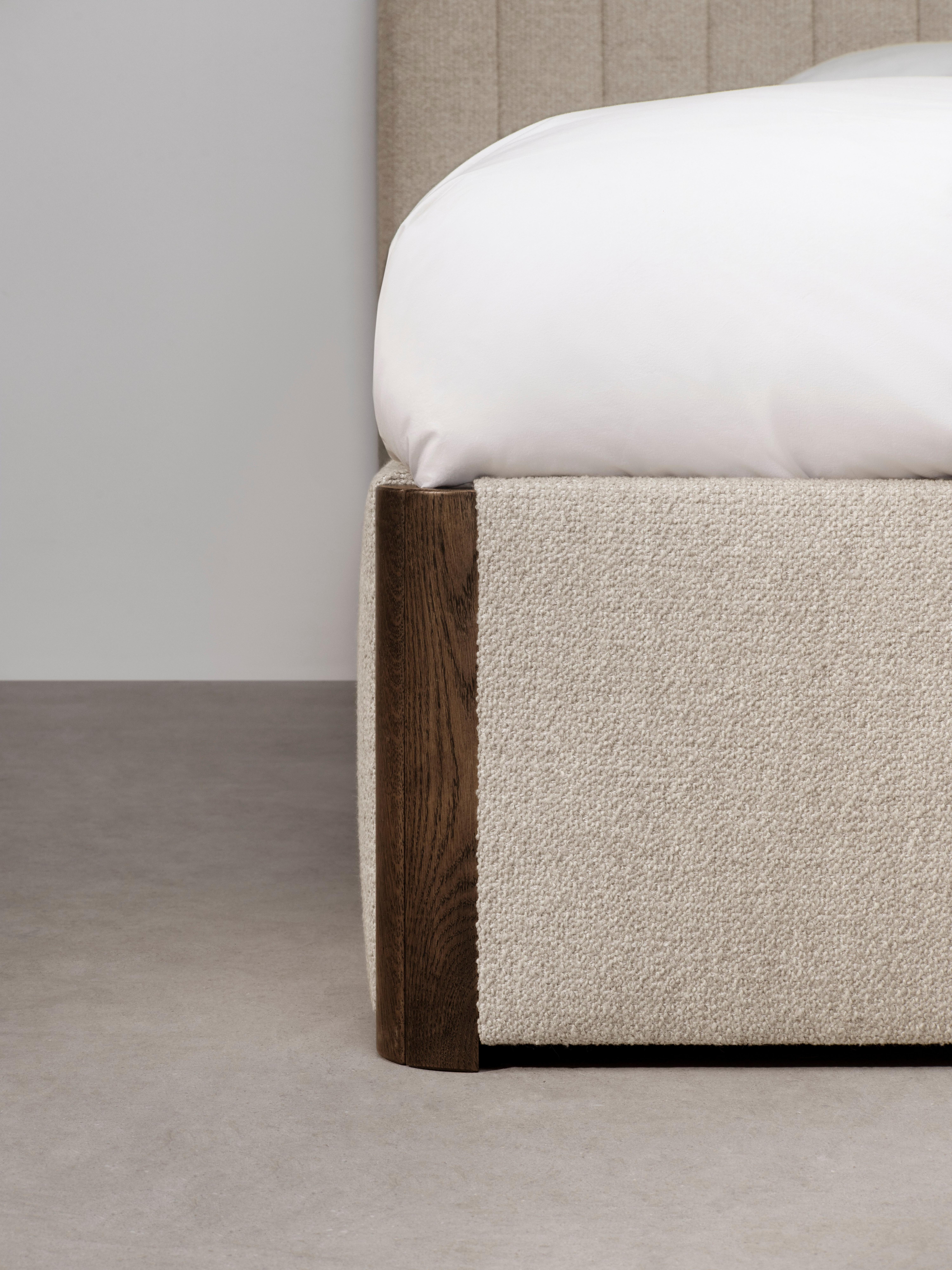 Beige upholstered bed corner with wooden frame and white bedding.