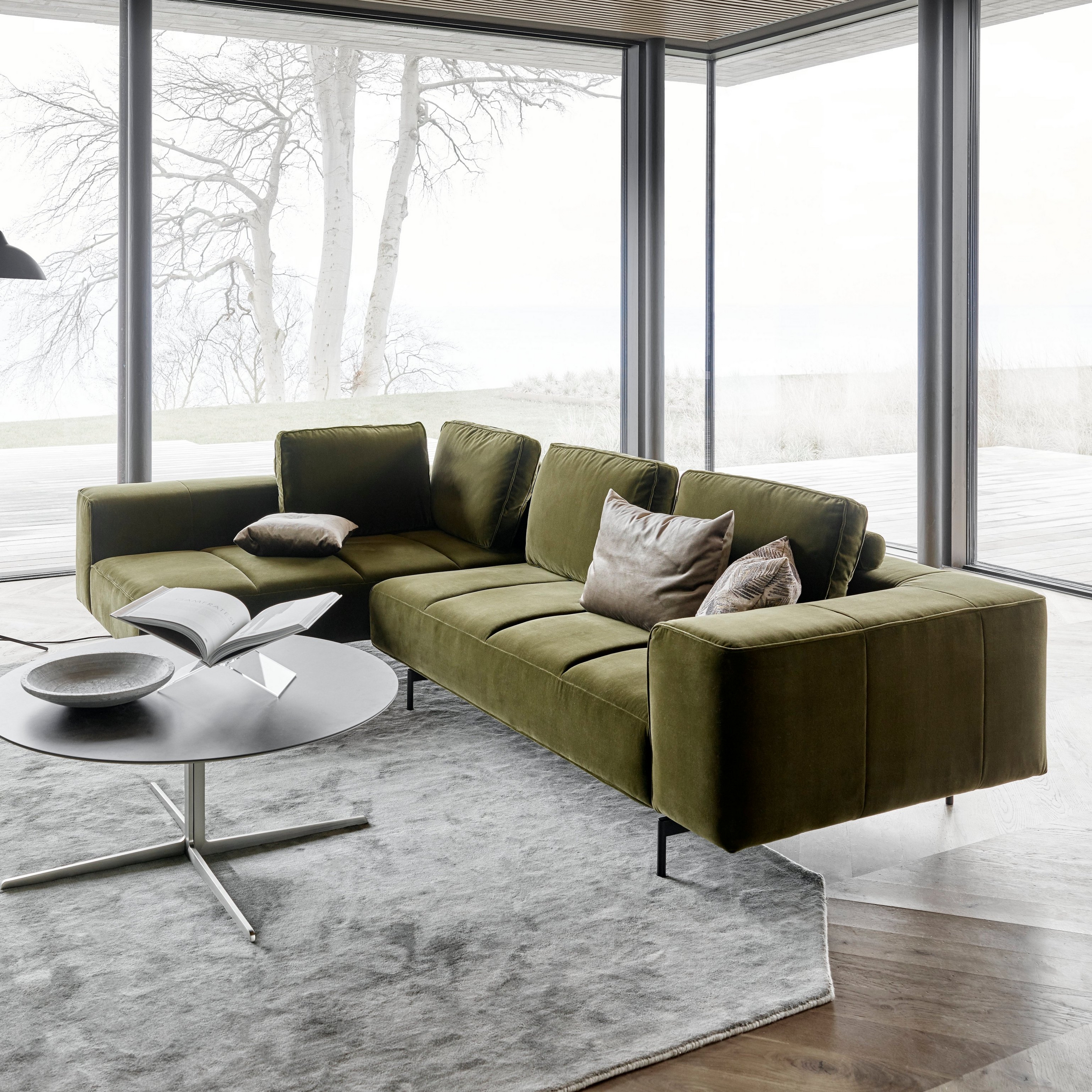 Modern grön Amsterdam soffa, soffbord, golvlampa som står vid ett panoramafönster med vattenutsikt.