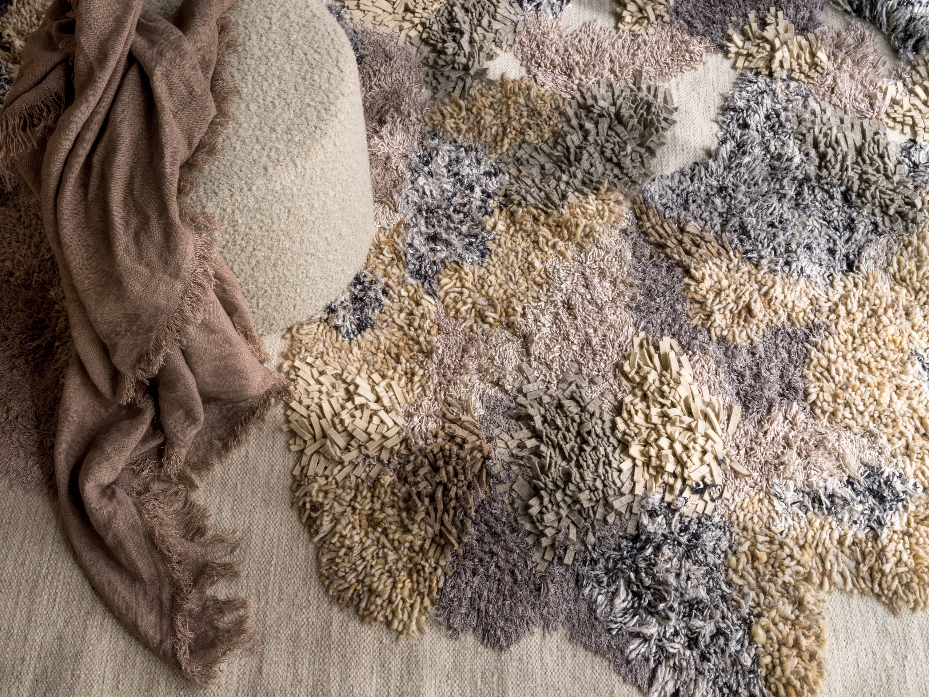 Primer plano de una alfombra Vivod texturizada con diseño abstracto y una bufanda drapeada sobre el puf Eden.