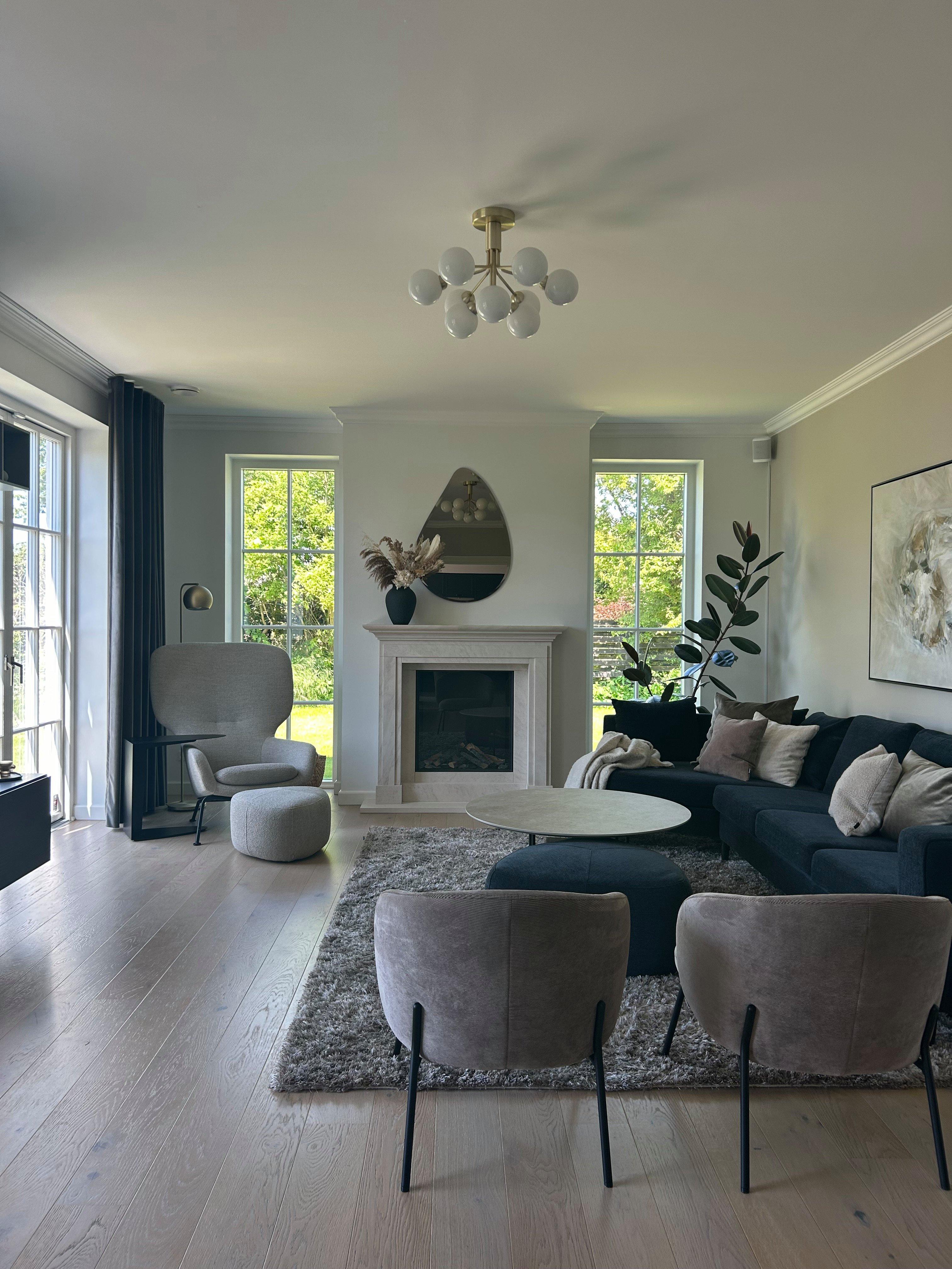 Modern living room with a neutral palette, featuring a dark sectional sofa, light accent chairs, and a central fireplace.