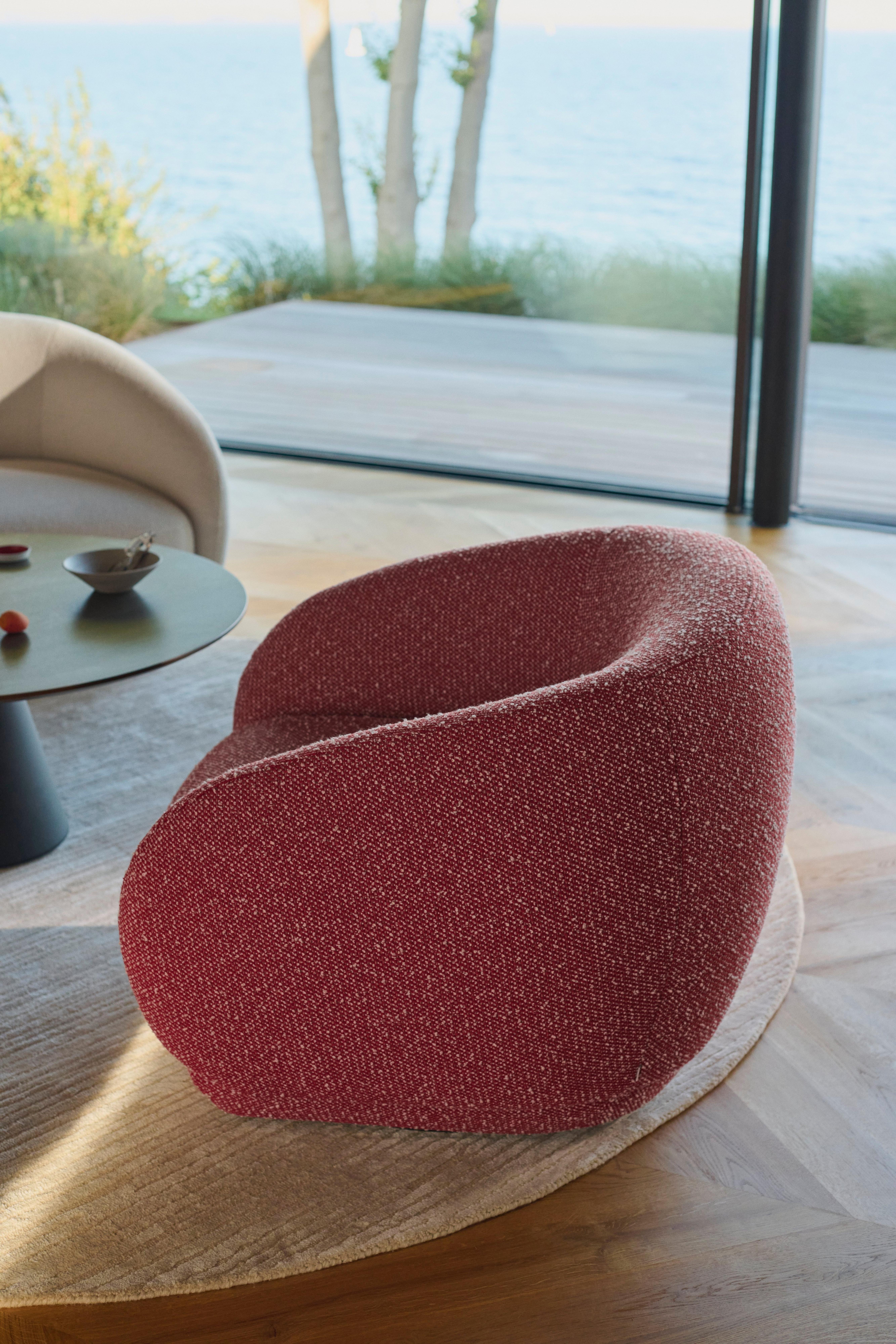 Nowoczesny fotel w czerwonej tkaninie o zakrzywionym kształcie na okrągłym beżowym dywanie, ustawiony przy szklanej ścianie z widokiem na morze.