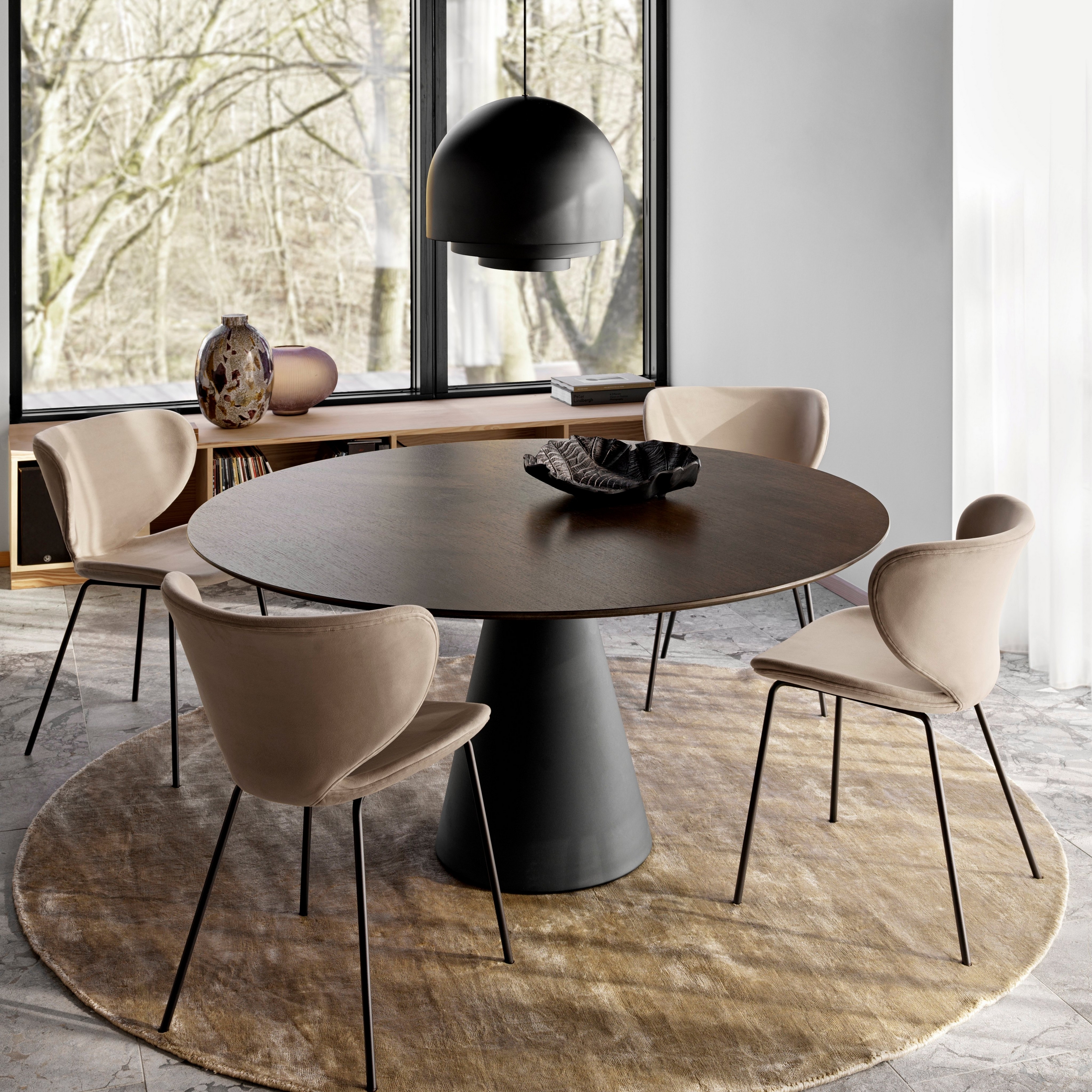 Runt matbord med bruna stolar på en rund matta nära fönster som sträcker sig från golv till tak, och en stor taklampa
