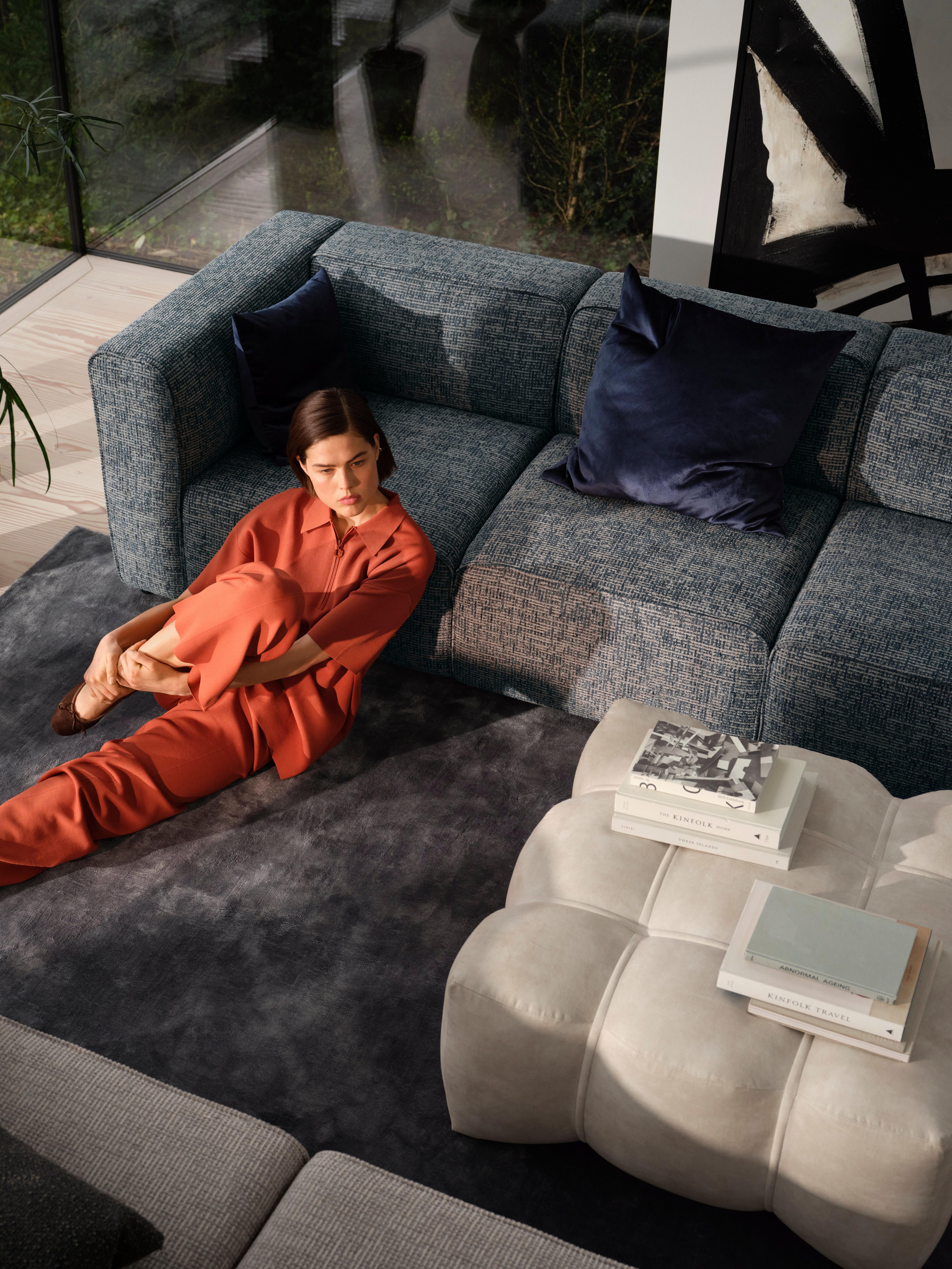 Living room featuring a woman in a rust-colored outfit sitting on a dark grey Loom rug next to a Nawabari footstool in beige Ravello fabric. The room includes a dark blue fabric sofa with dark blue velvet cushions, white Tuscany fabric, and abstract black and white wall art. Large windows provide an outdoor view.