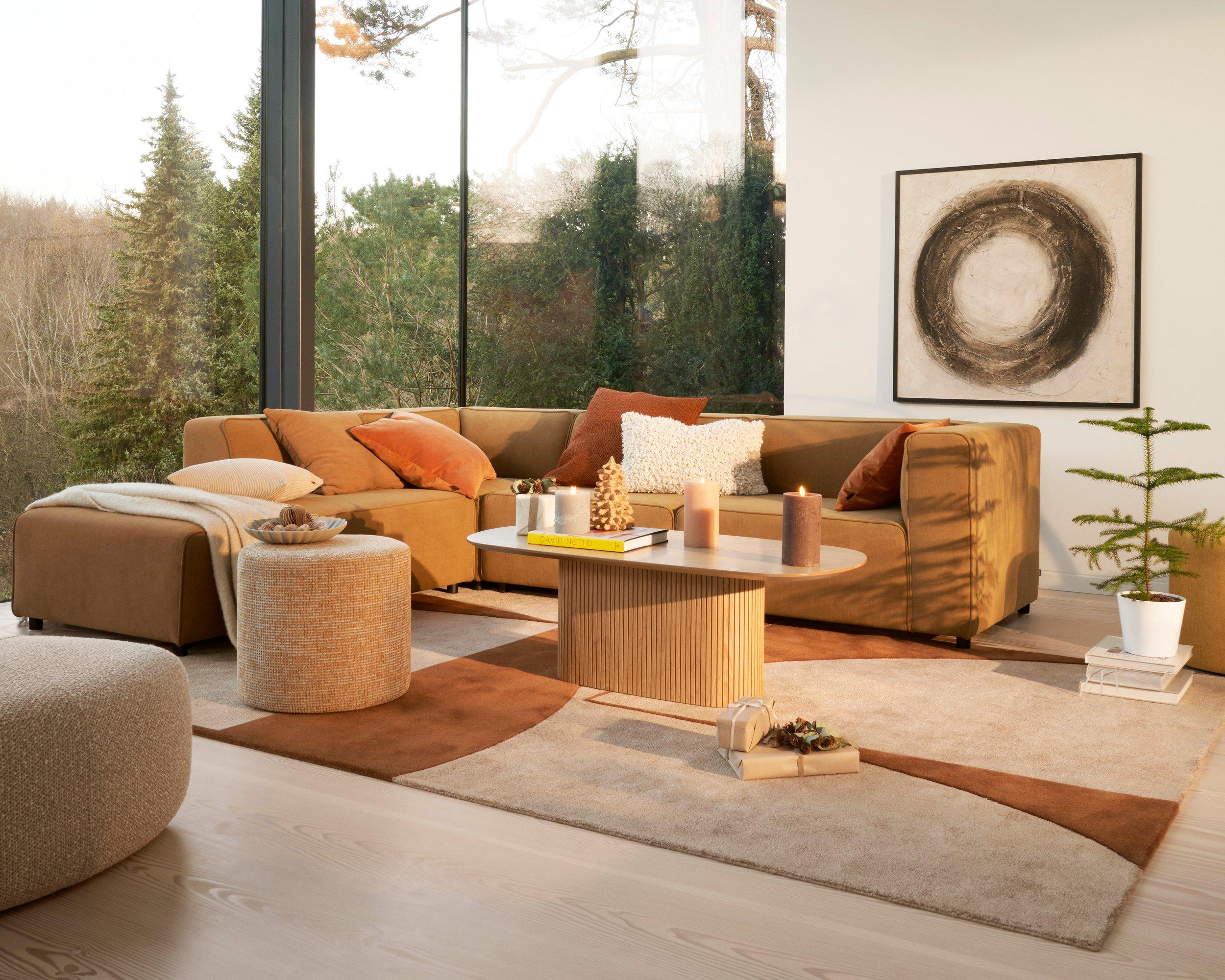 Salon moderne avec un canapé sectionnel Carmo camel avec coussins rouille, un repose-pieds Eden rond beige et une table basse Santiago en bois clair.