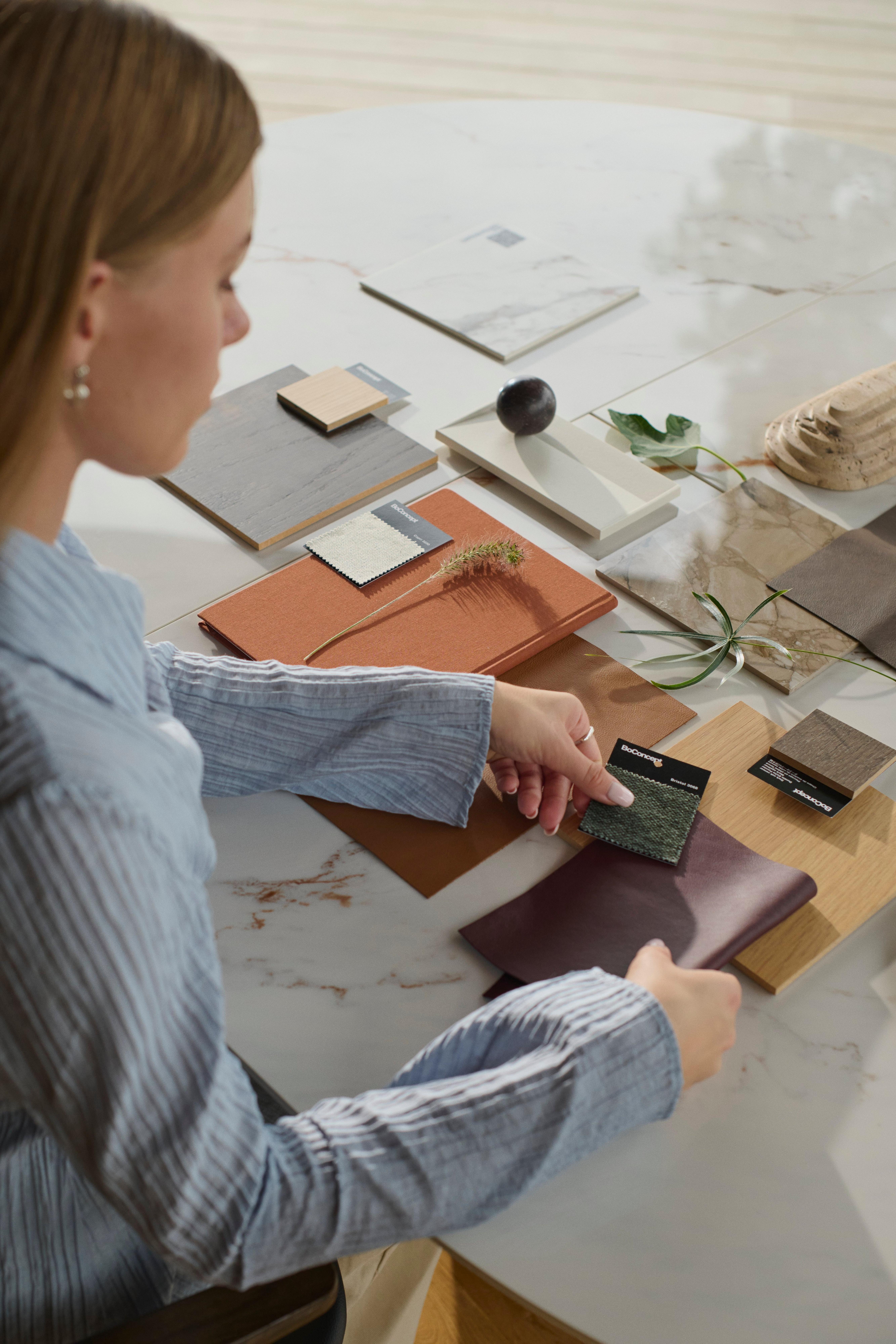 A woman selects fabric and material samples on a marble table, arranging wood, textiles, and stone finishes in a neutral-toned palette.