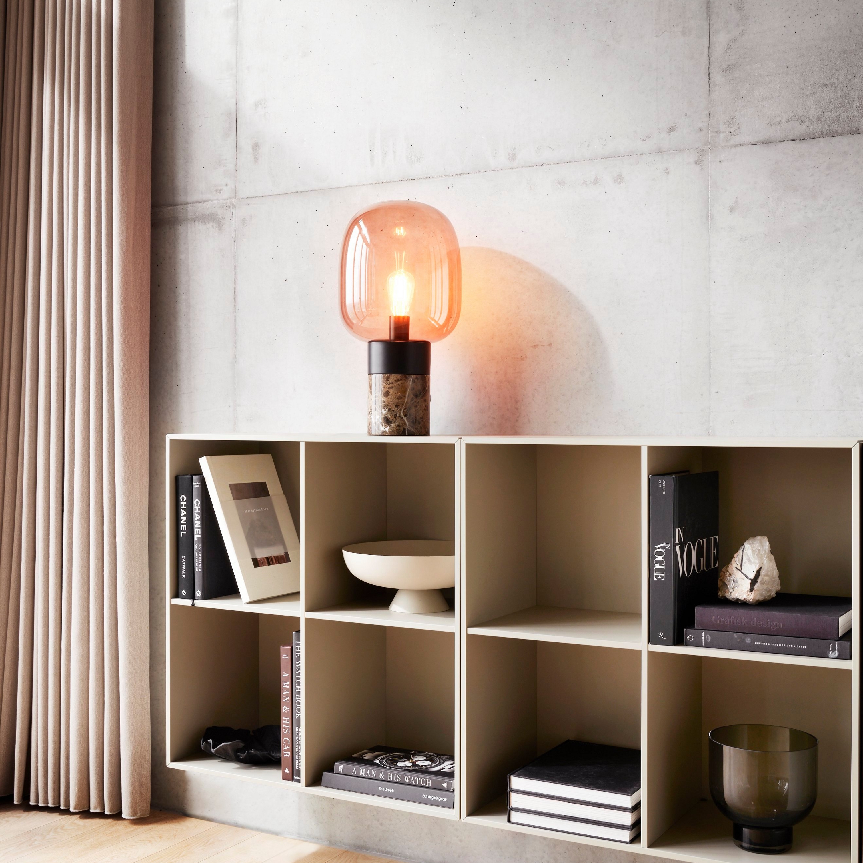 Modern shelf with books, bowl, and lamp against a concrete wall, with curtains to the side.