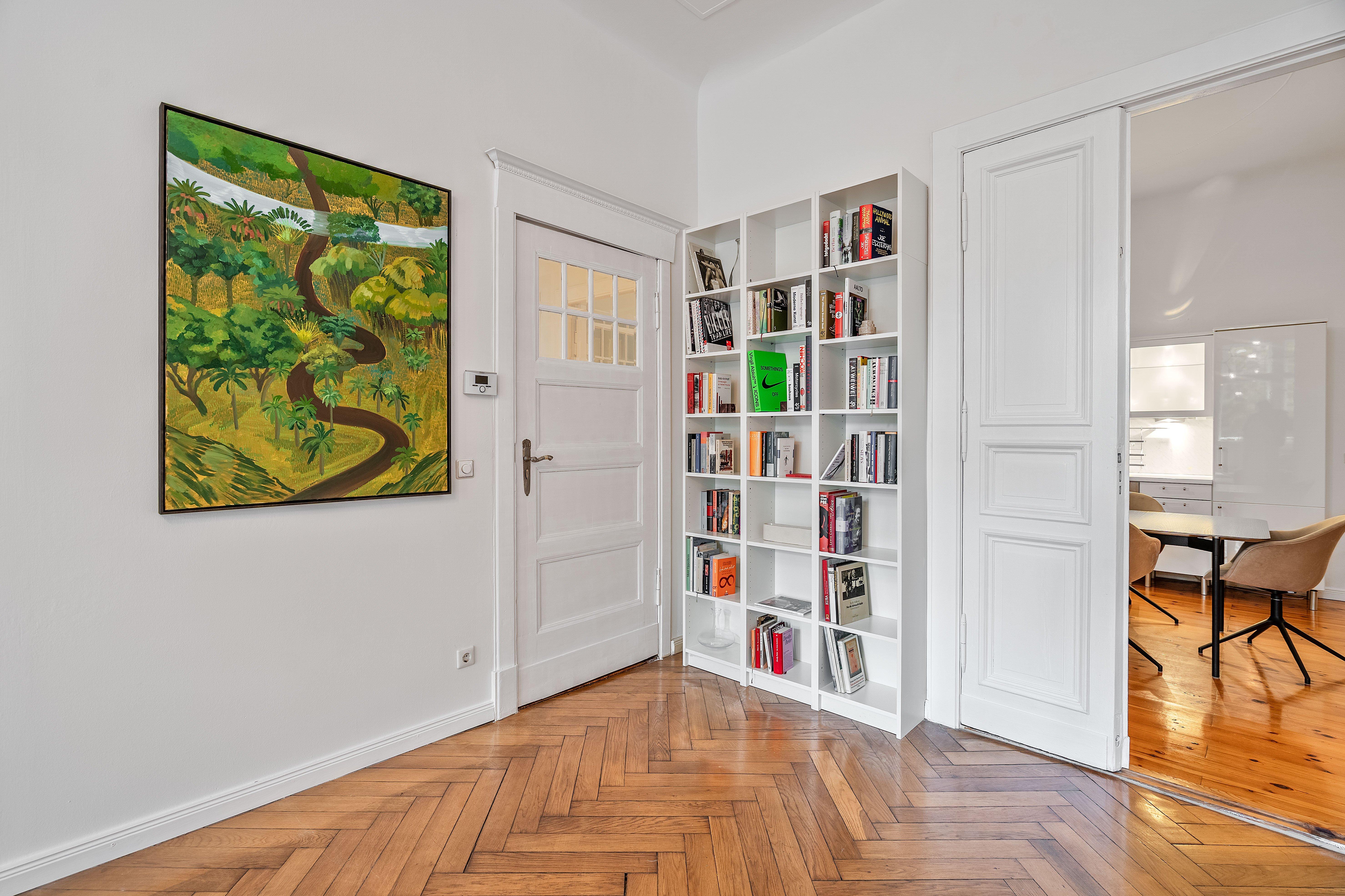 Tableau coloré de paysage, étagère avec livres, porte blanche ouverte sur une salle à manger.