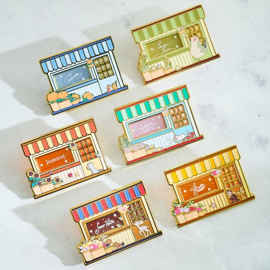 Six colorful Disney princess enamel pins shaped like storefronts with awnings and illustrated animals. Shop Now.