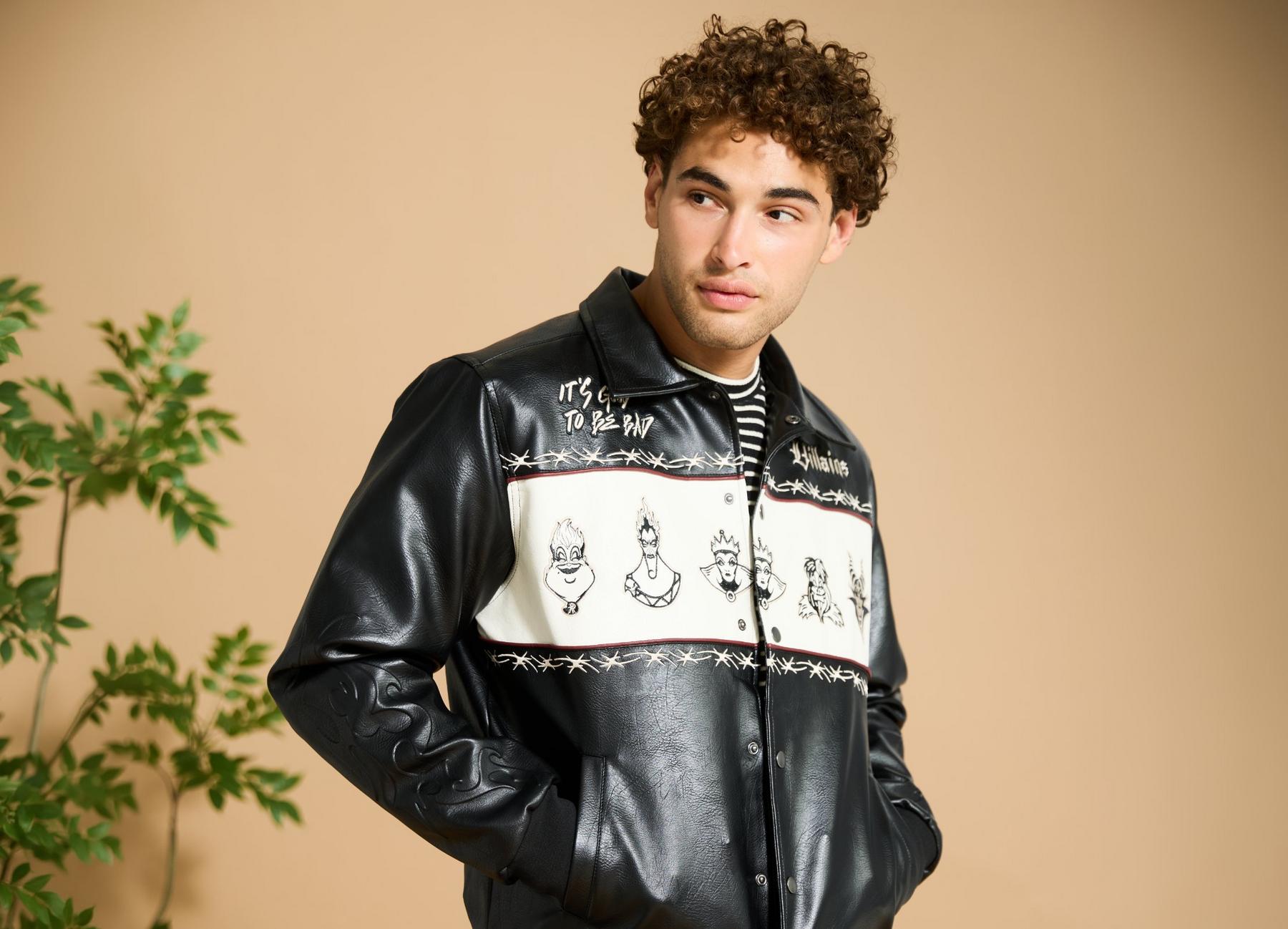 Man modeling black faux leather jacket with white panel and villain sketches, hands in pockets. Shop Now.