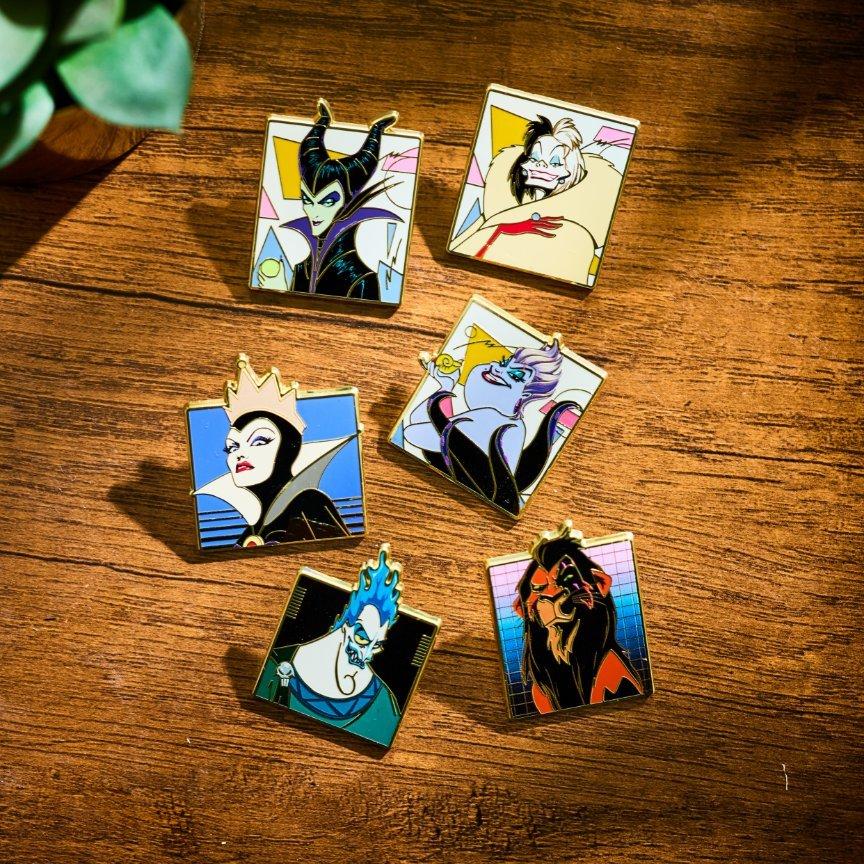 Six Disney Villains character enamel pins on a wooden surface. Shop Now.