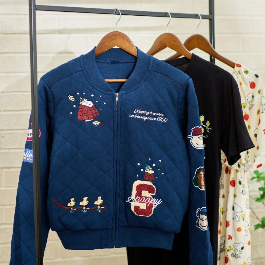 Quilted blue zip-up jacket with Snoopy and Woodstock patches hanging on a rack with other Snoopy and Woodstock styles. Shop Now.
