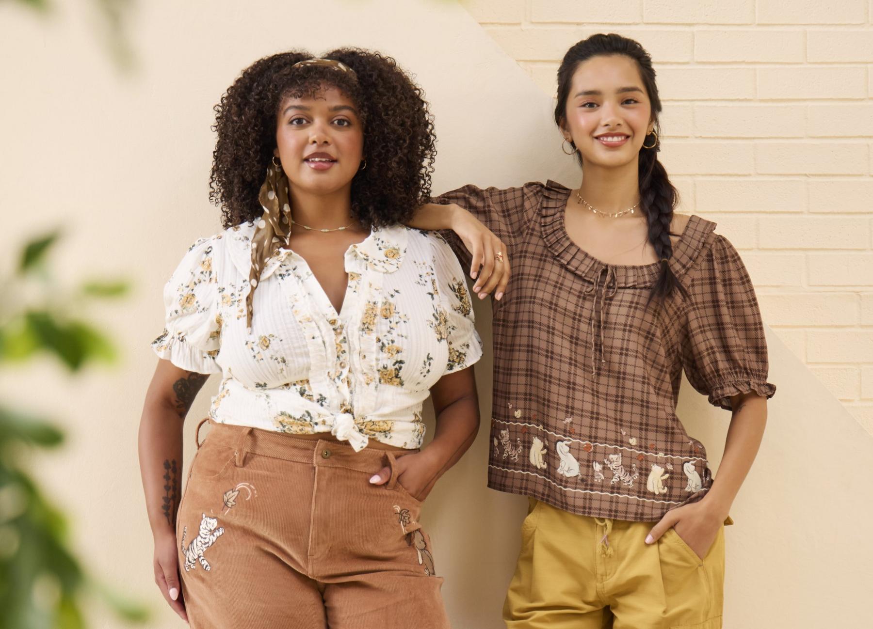 Two women modeling a floral blouse and brown embroidered Winnie the Pooh pants, and a brown plaid Winnie the Pooh blouse with yellow pants. Shop Now.