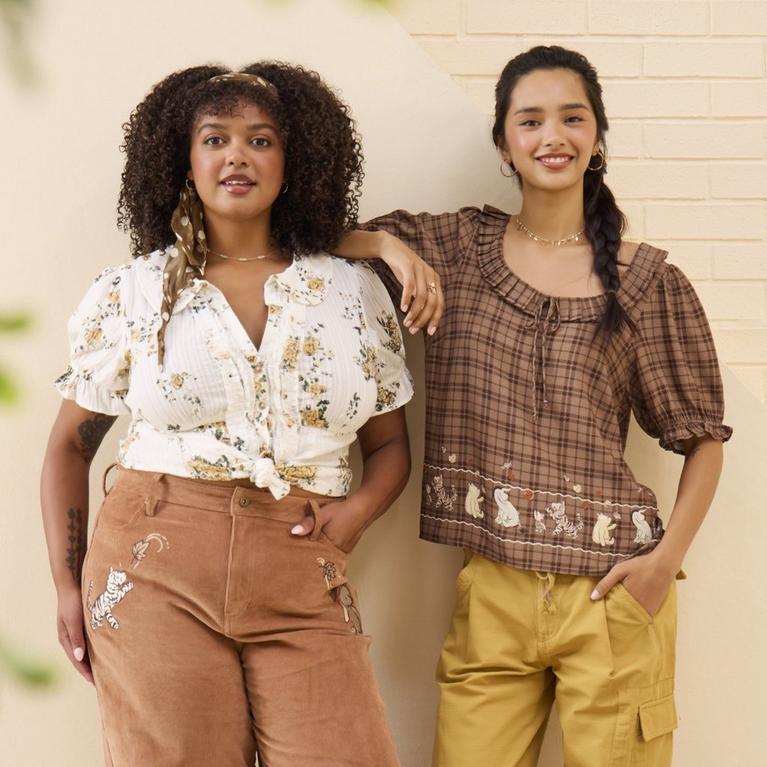 Two women modeling a floral blouse and brown embroidered Winnie the Pooh pants, and a brown plaid Winnie the Pooh blouse with yellow pants. Shop Now.