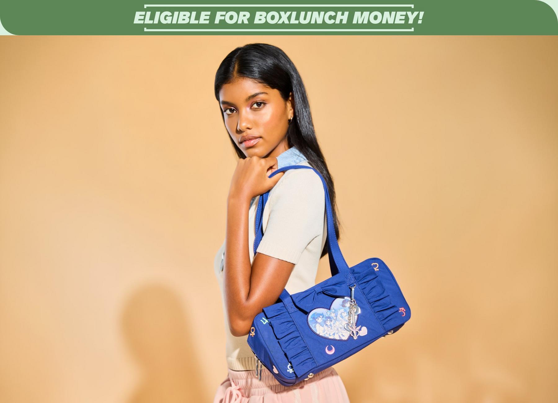 Model holding a blue Sailor Moon shoulder bag with a heart graphic patch and dangling charm under “ELIGIBLE FOR BOXLUNCH MONEY!” banner. Shop Now.