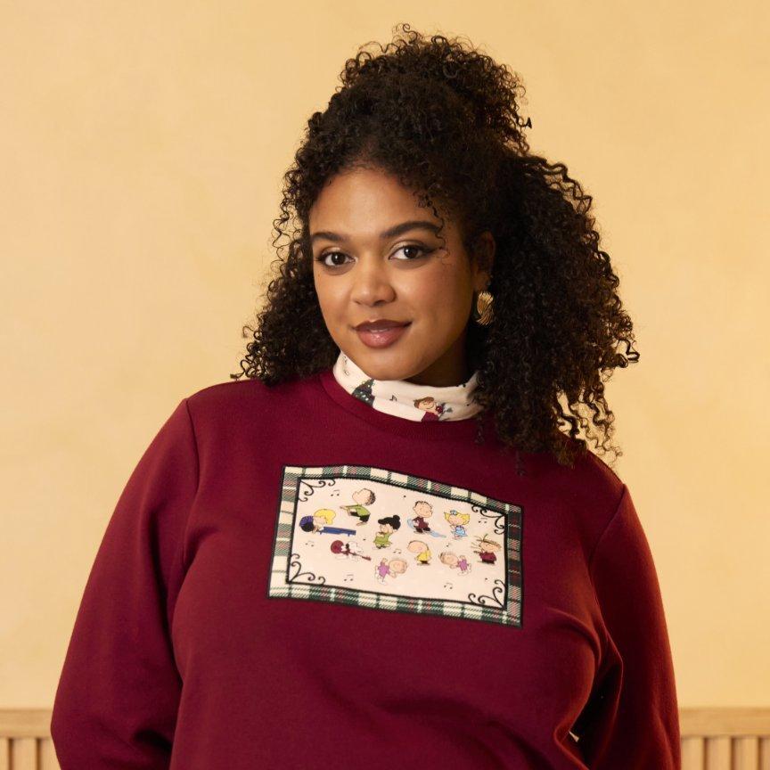 Model wearing a burgundy sweatshirt with cartoon graphic patch of the Peanuts friends and a printed turtleneck underneath. Shop Now.