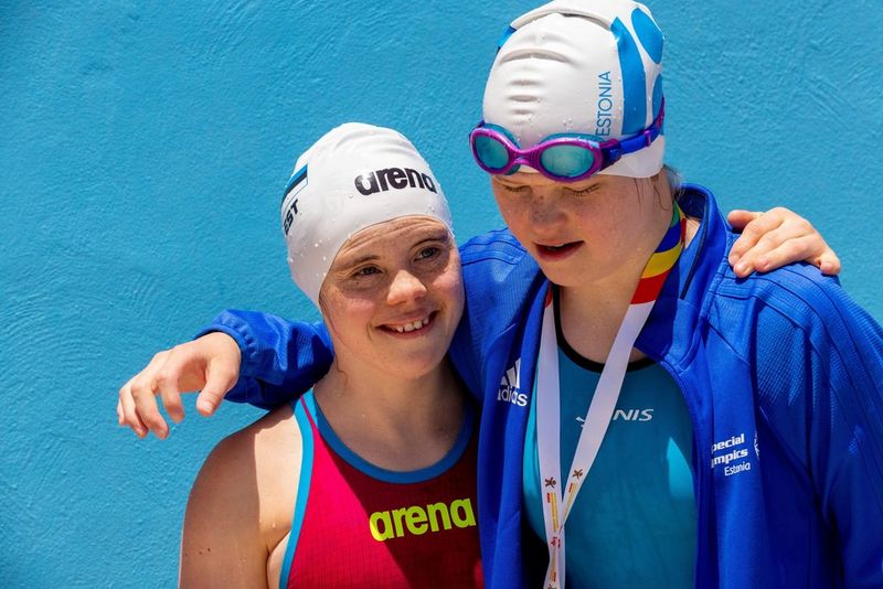 Two swimmers photographed from the chest up. The swimmer on the right has their arm around the shoulders of the swimmer on the left. They are both wearing white swimming caps, with goggles pushed up over it on the right-hand swimmer. She also wears a blue jacket over a blue swimming costume and the ribbon of a medal can be seen around her neck. The left-hand swimmer wears a red costume with blue trim.