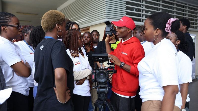 Tore Oriase, in een rode Canon-hoodie en bijpassende pet, demonstreert een professionele Canon-videocamera aan een groep studenten.