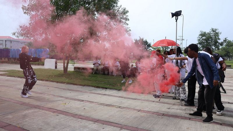 White Man draagt een donker jasje en een broek met patroon en staat midden in een wolk roze rook die op hem wordt gericht door een groep studenten aan de rechterkant. De studenten filmen hem terwijl hij beweegt.
