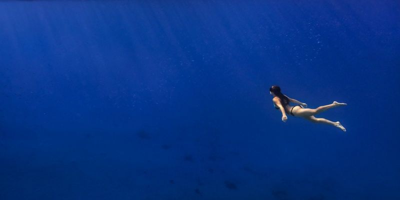 An image taken underwater. The sea is bright blue and a woman in a bikini and snorkel swims upwards towards the light and bubbles on the surface. © Dafna Tal