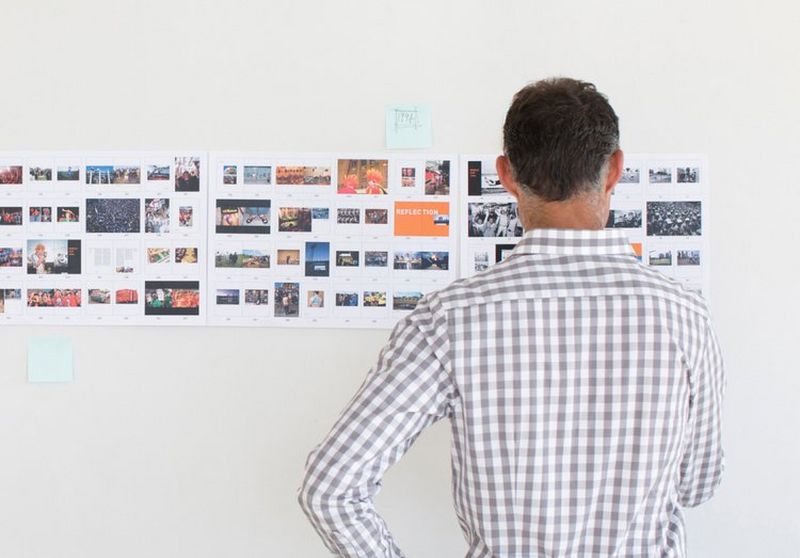Man in a checked shirt with his back towards the camera stands with his hand on his hip as he stares at tiny images spread across a white wall.