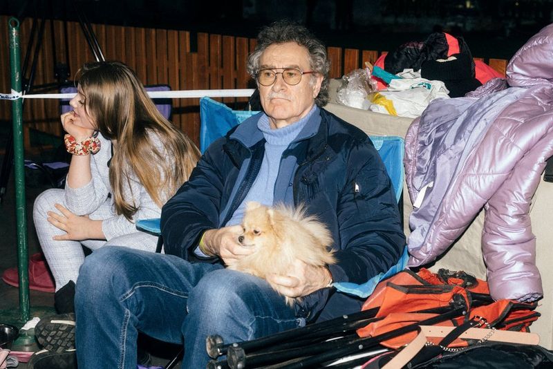 A man sat next to a girl holding a miniature spitz dog in his lap amongst a puffer coat and folded orange deckchair. Shot on Canon.