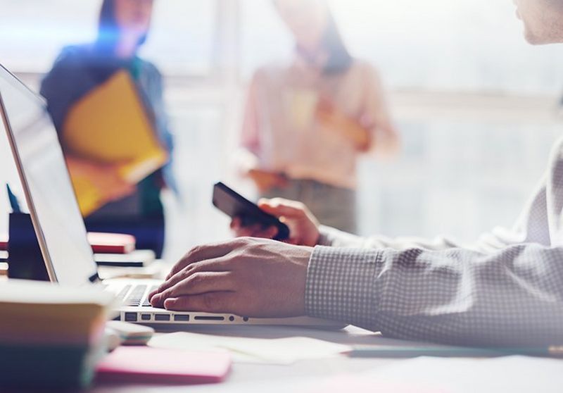 Man typing on laptop whilst holding his phone up