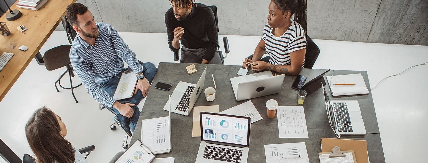 Overhead shot of casually dressed office workers sat around a communal table comparing graphs on laptops and clipboards.