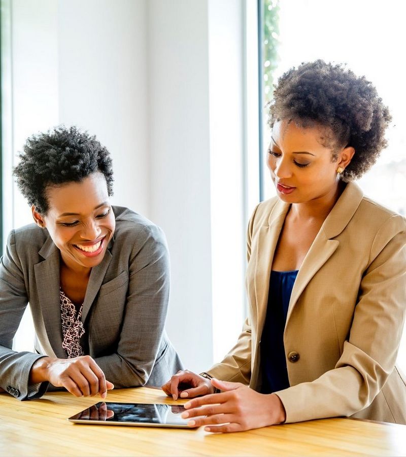 Two women dressed in jackets smile as they look at something on a tablet device laid flat on a desk.