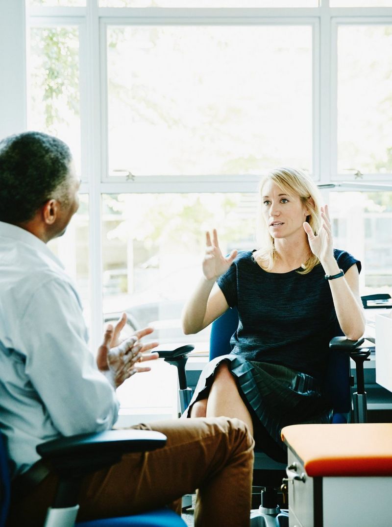 Woman dressed in black sits in front of a window and gestures with her hands to a man in beige trousers sitting across from her.
